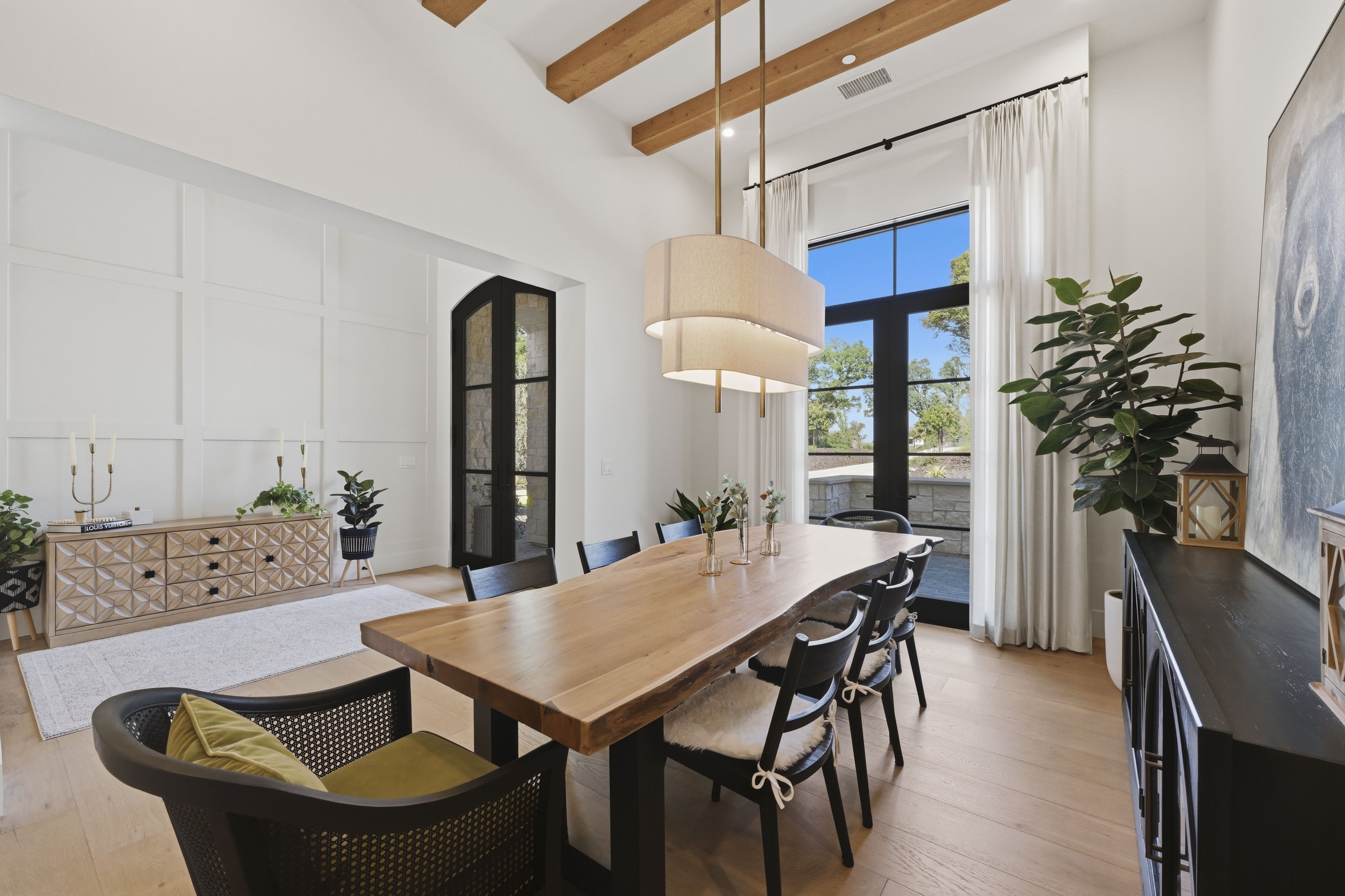 Interior of a modern dining room with high vaulted ceiling featuring wooden beams, large window with white curtains, and a wooden dining table surrounded by black chairs. A large potted plant is near the window, and a contemporary chandelier hangs ov