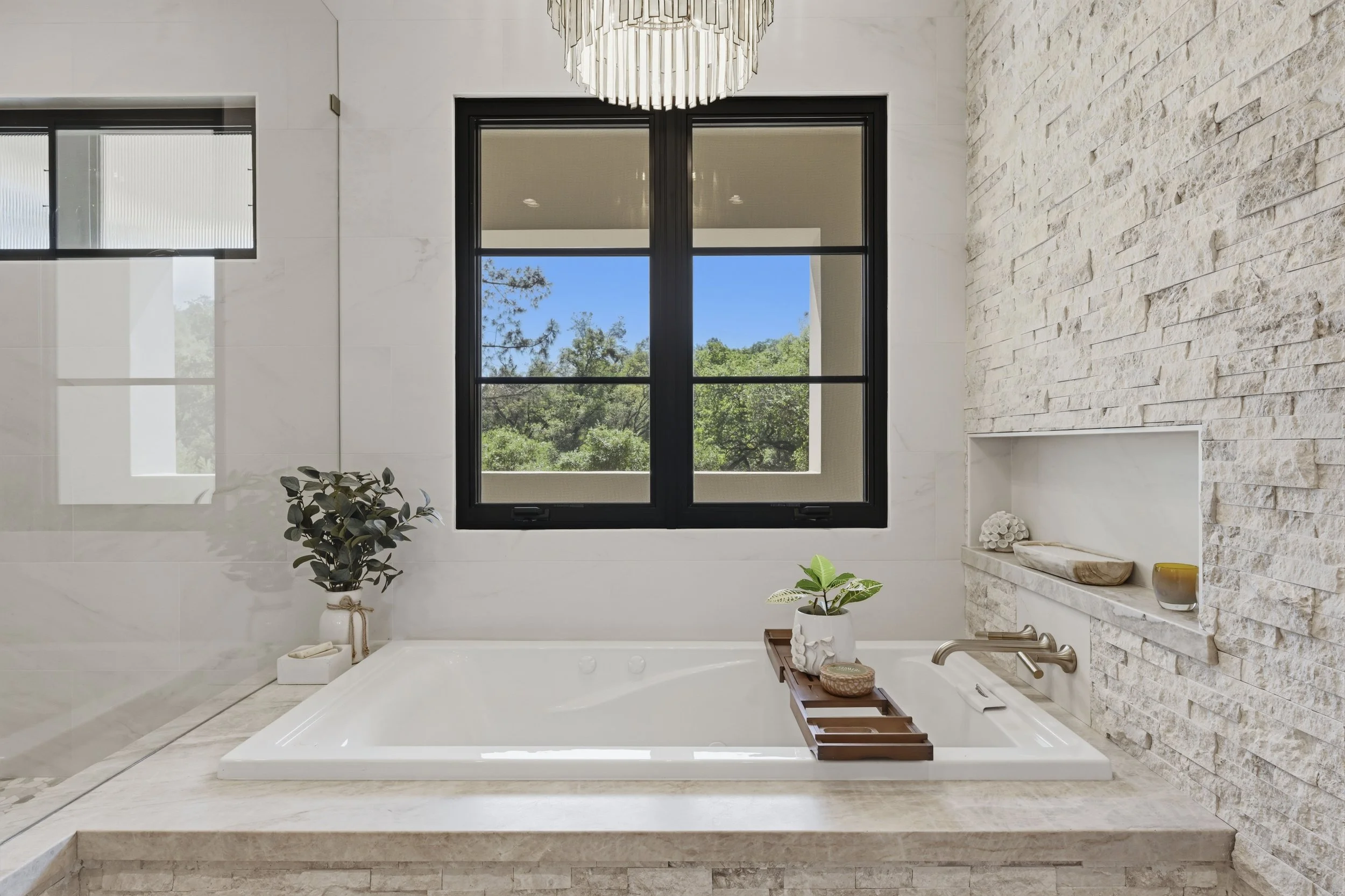 Bathroom with large window showing trees and blue sky, white stone wall, potted plants, and a bathtub with decorative tray and candles.