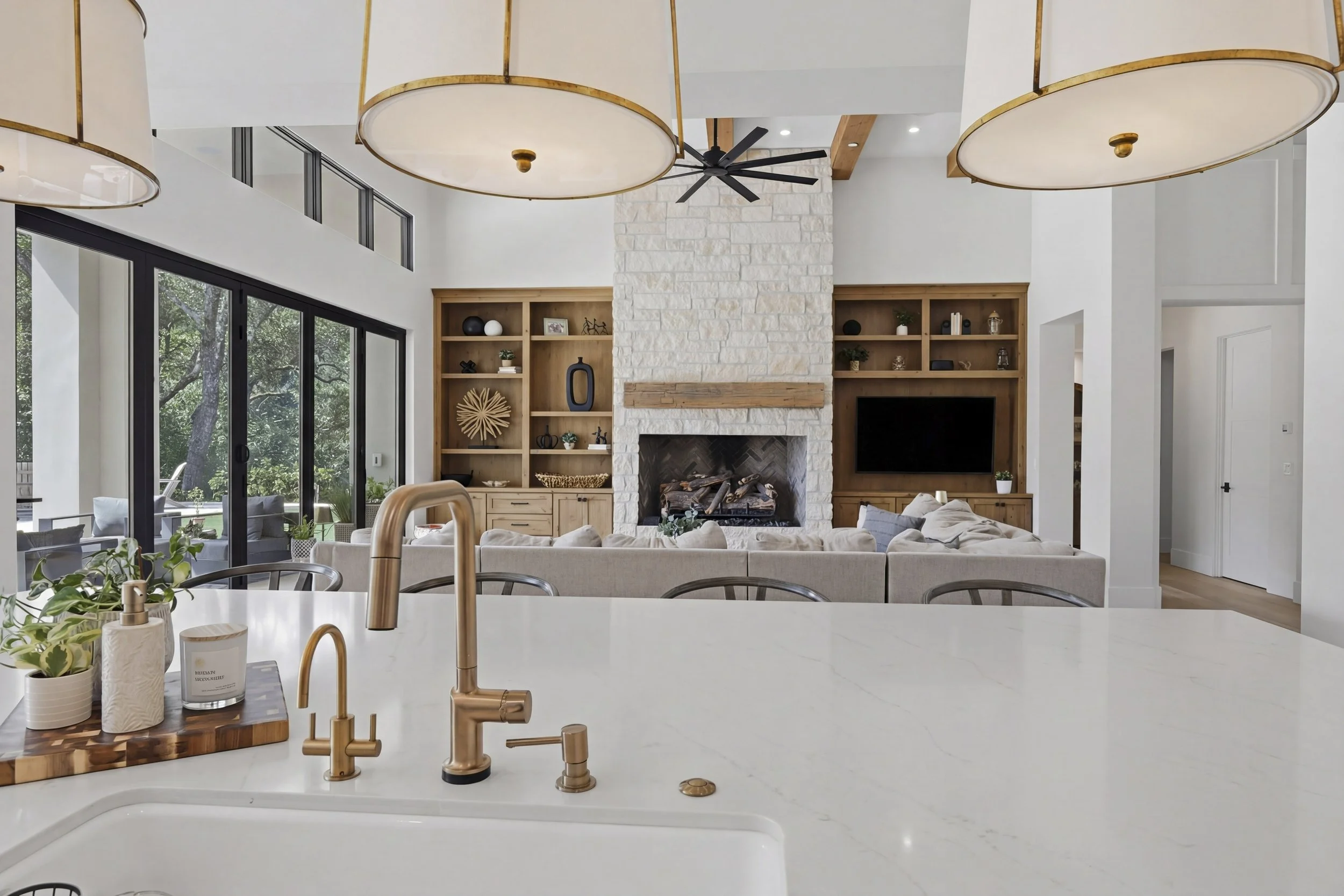 A modern open-concept living room and kitchen with large windows showing outdoor trees. The room features a white stone fireplace, built-in wooden shelves, a wall-mounted flat-screen TV, and a ceiling fan. The kitchen has a white countertop with a br