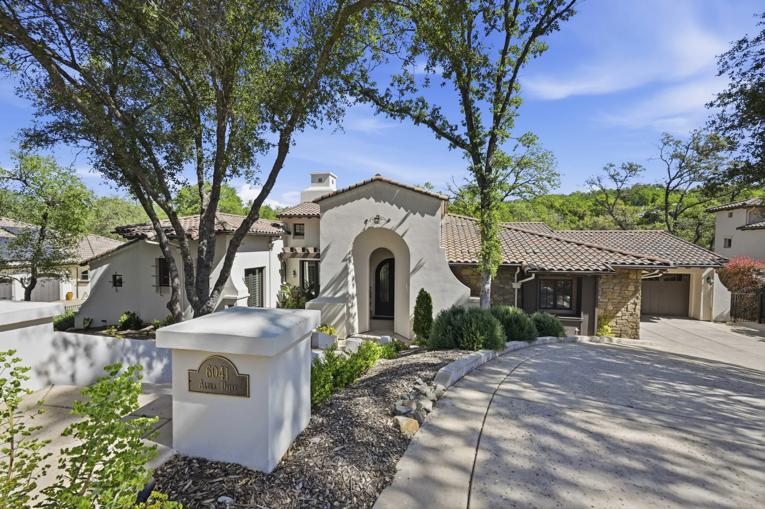 A modern house with white walls, tile roof, surrounded by trees and greenery, with a curved driveway leading to the entrance and a mailbox that displays the address 6041 Aldea Drive.