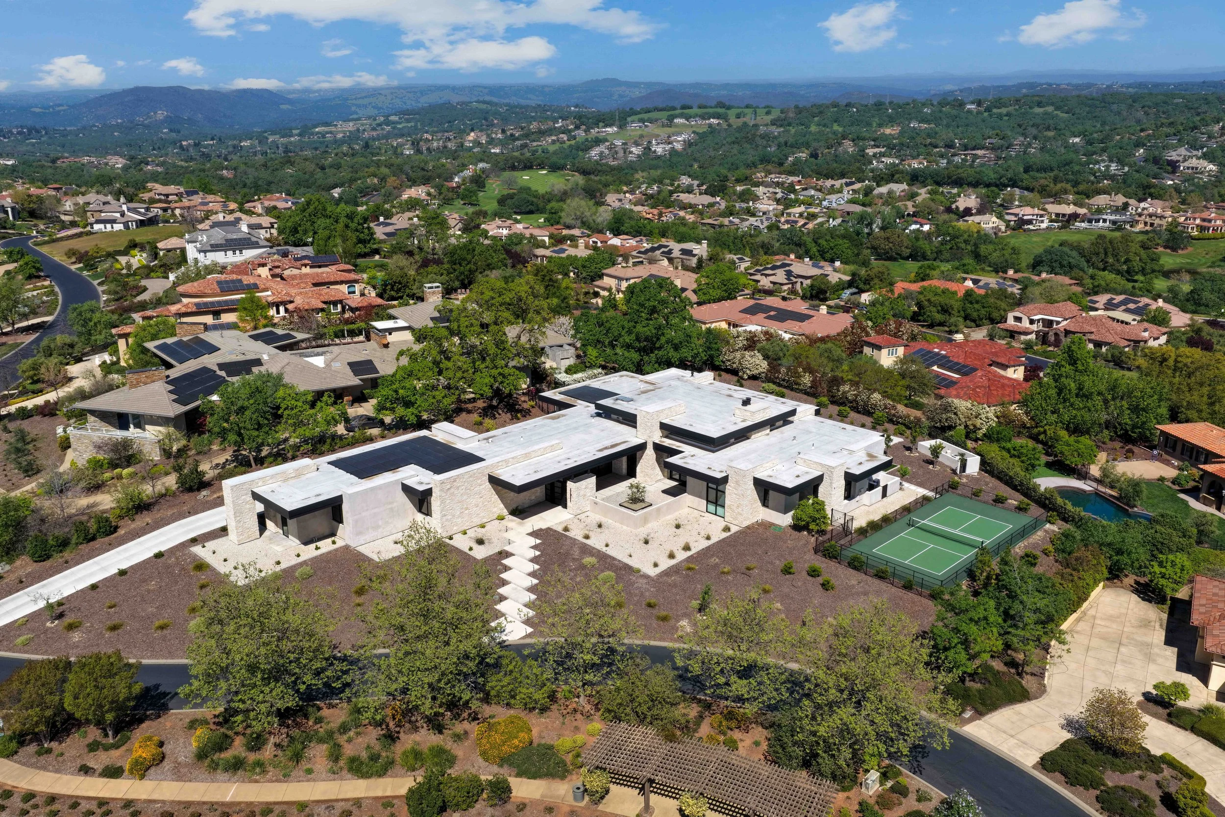 Aerial view of a suburban neighborhood with houses, greenery, and distant mountains under a partly cloudy sky.