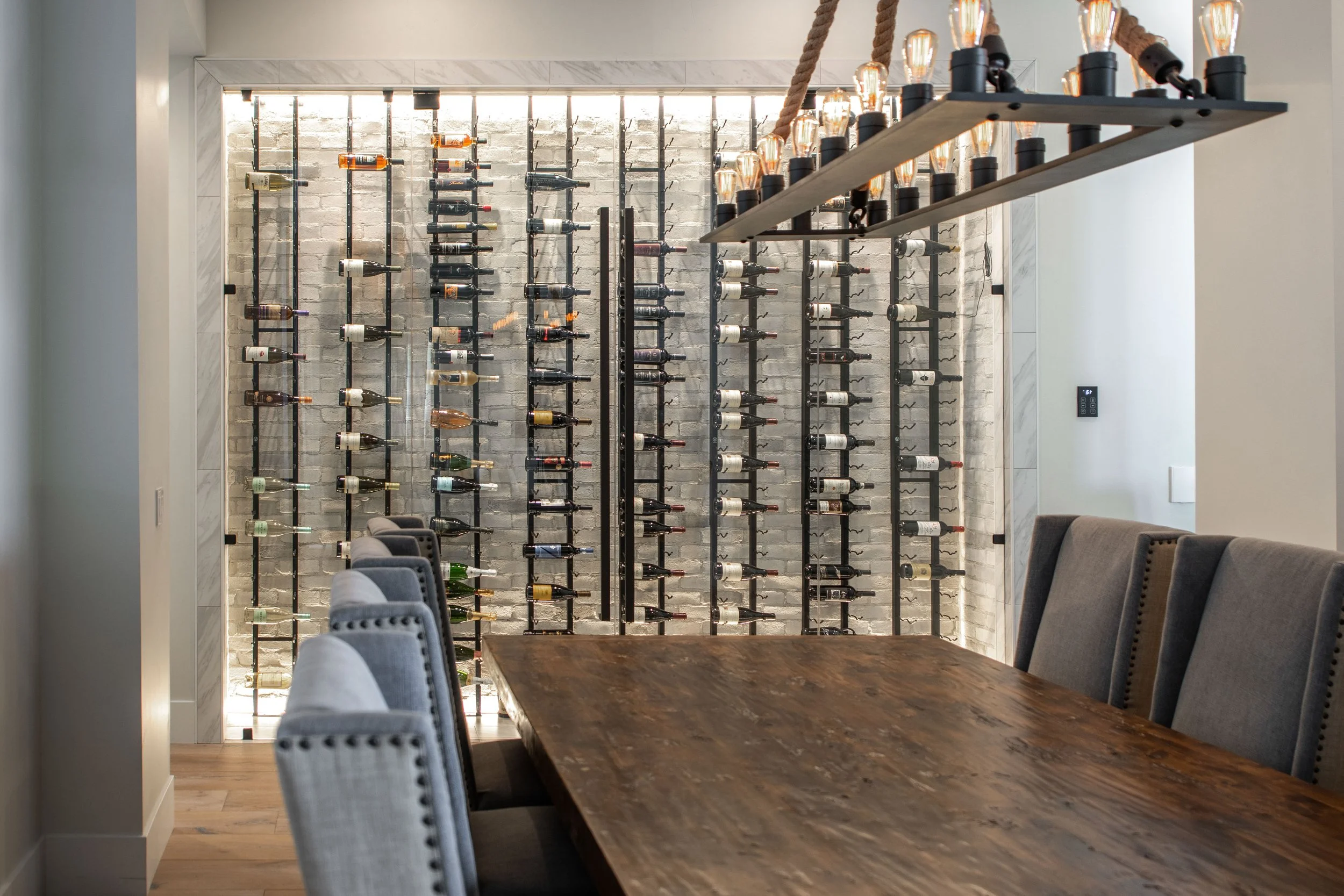 A modern dining room with a large wooden table and upholstered chairs. In the background, there is a wall display of wine bottles on metal racks against a white brick wall, and a hanging light fixture with exposed bulbs.