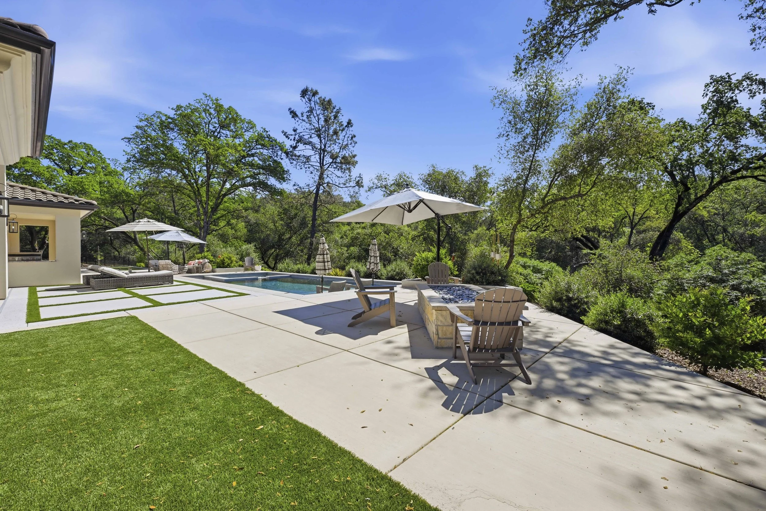 A backyard patio with a swimming pool, outdoor furniture, and umbrellas surrounded by trees and greenery under a blue sky.