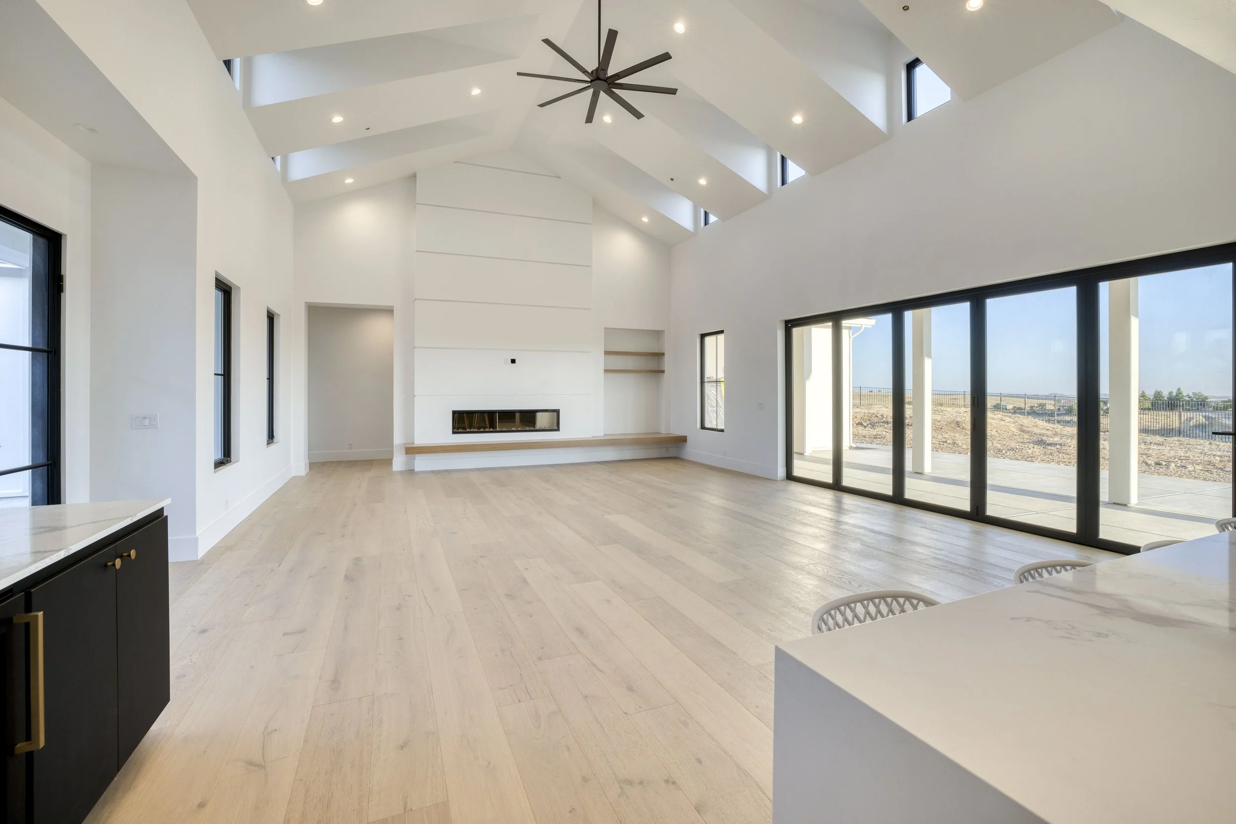 Modern living room with a vaulted ceiling, black ceiling fan, multiple windows allowing natural light, light wood flooring, and a large sliding glass door leading to an outdoor area.