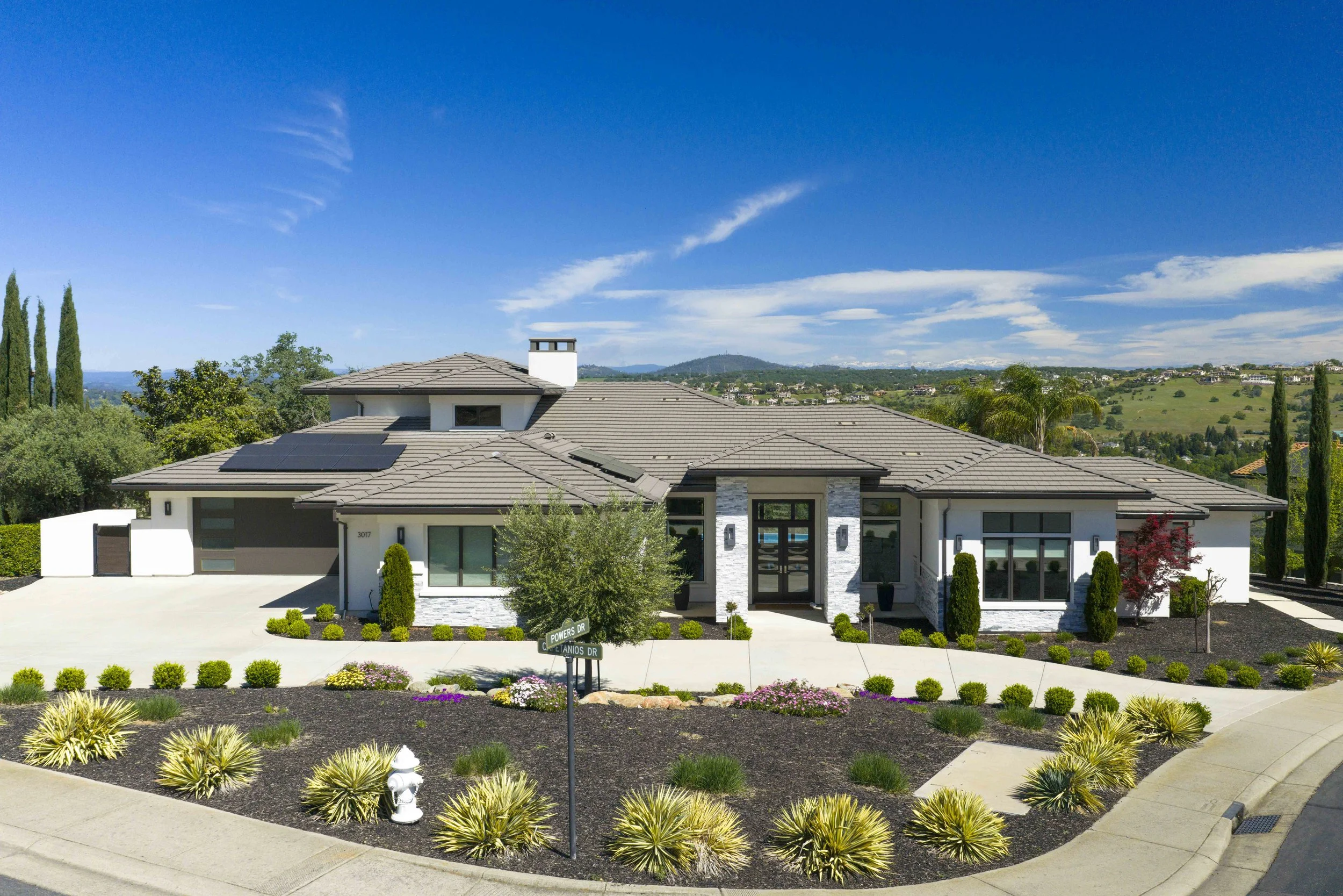 Modern white house with solar panels on the roof, surrounded by landscaped garden and trees, with hilly landscape and blue sky in the background.