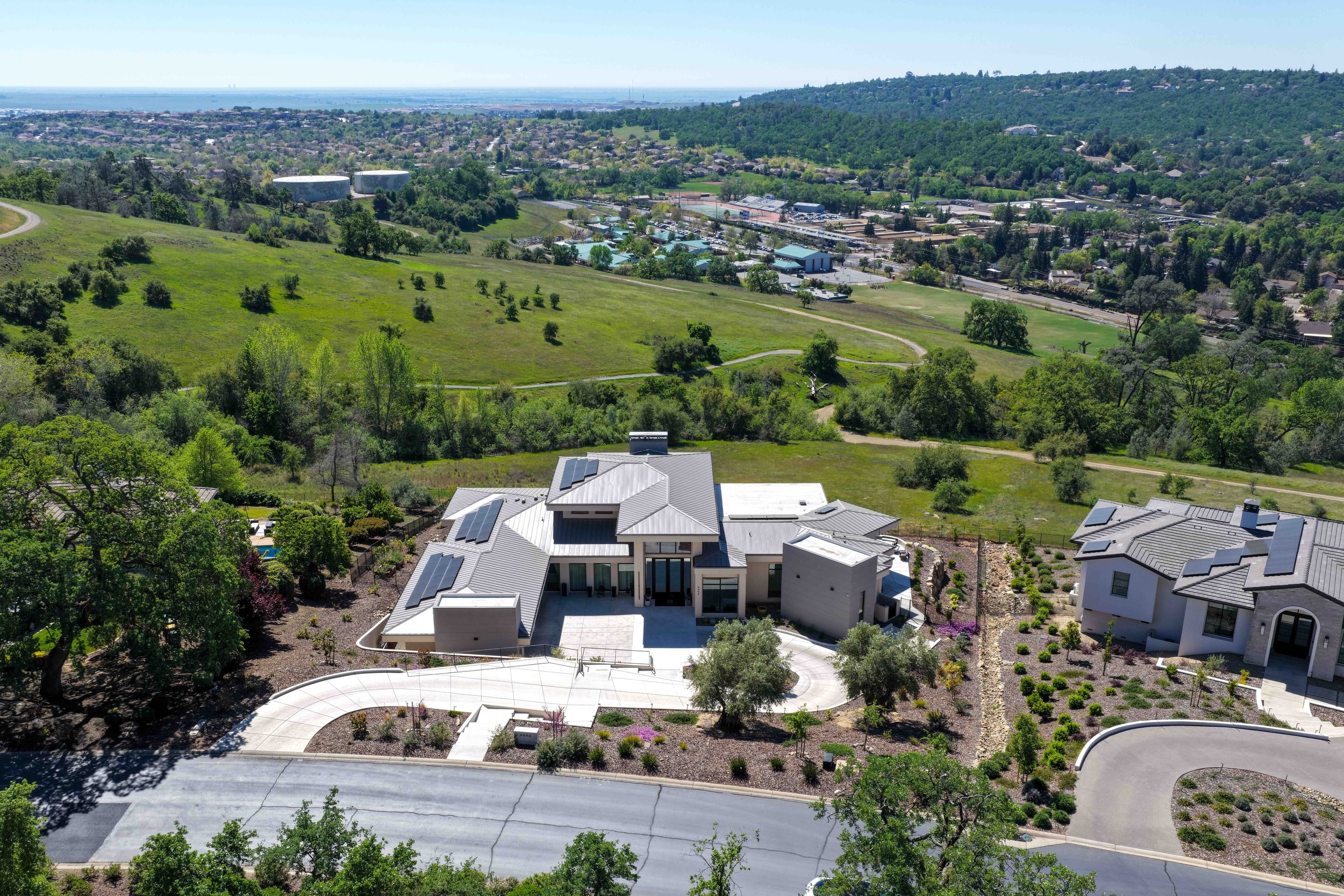 Aerial view of a modern house with solar panels, surrounded by landscaped garden, overlooking green hills, with a neighborhood and city in the distance.