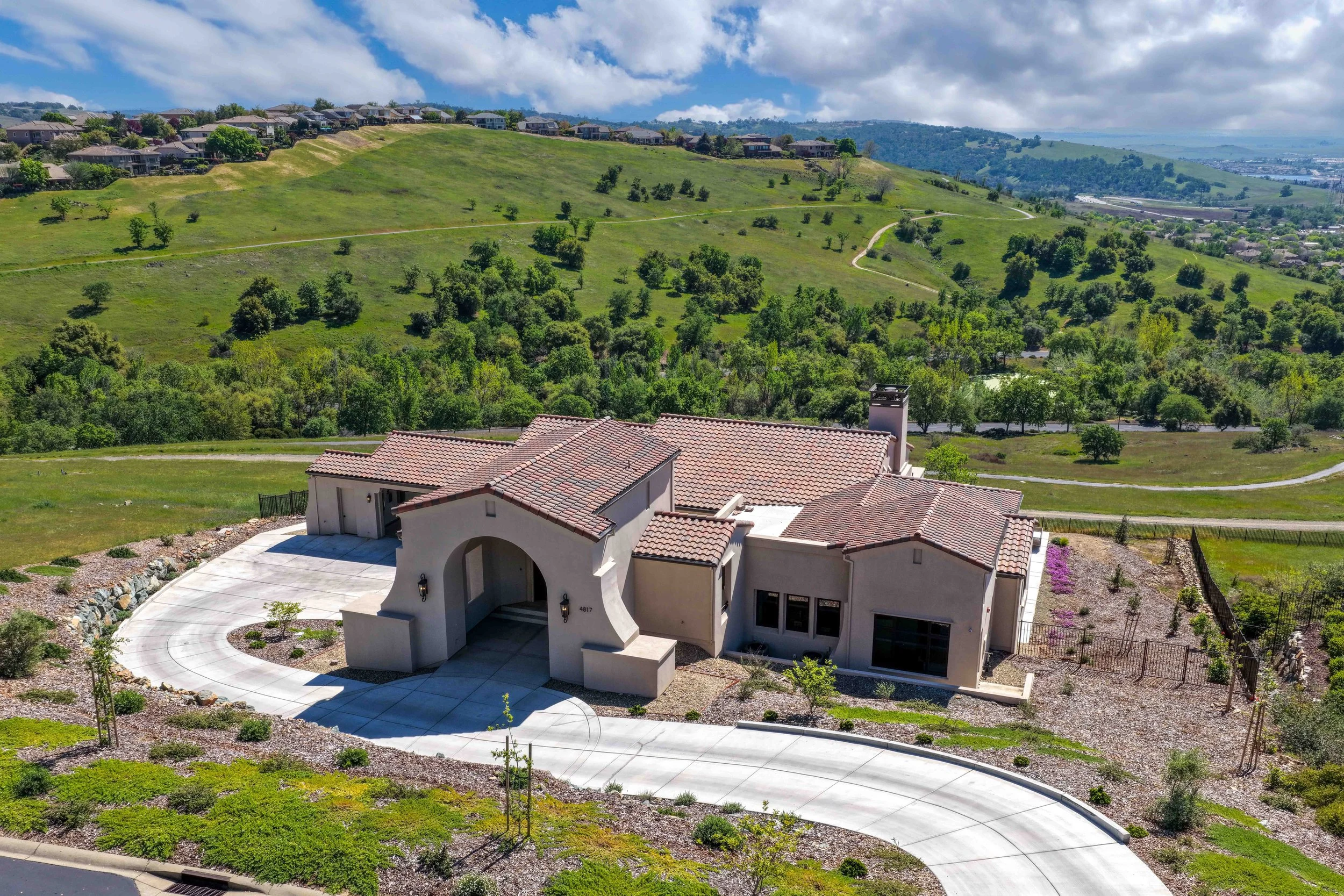 Aerial view of a suburban house with a red tile roof and a curved concrete driveway, set against green rolling hills and a partly cloudy sky.
