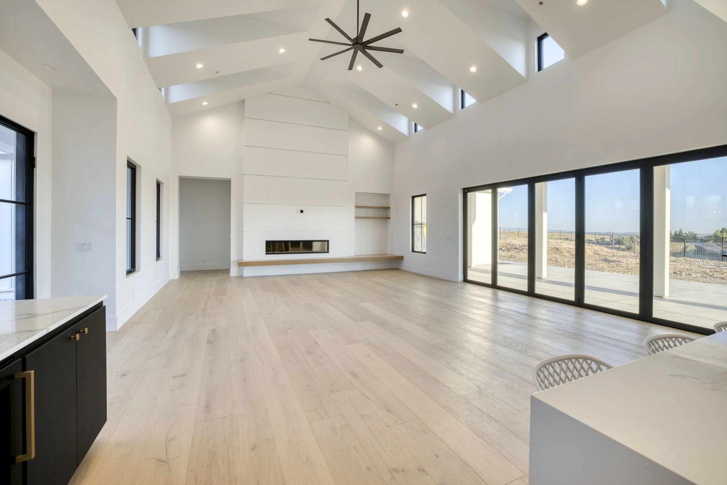 Interior of a spacious, modern living room with high white walls, large windows, and a vaulted ceiling with a ceiling fan. The room features light wood flooring, a fireplace, and a view of a patio outside.