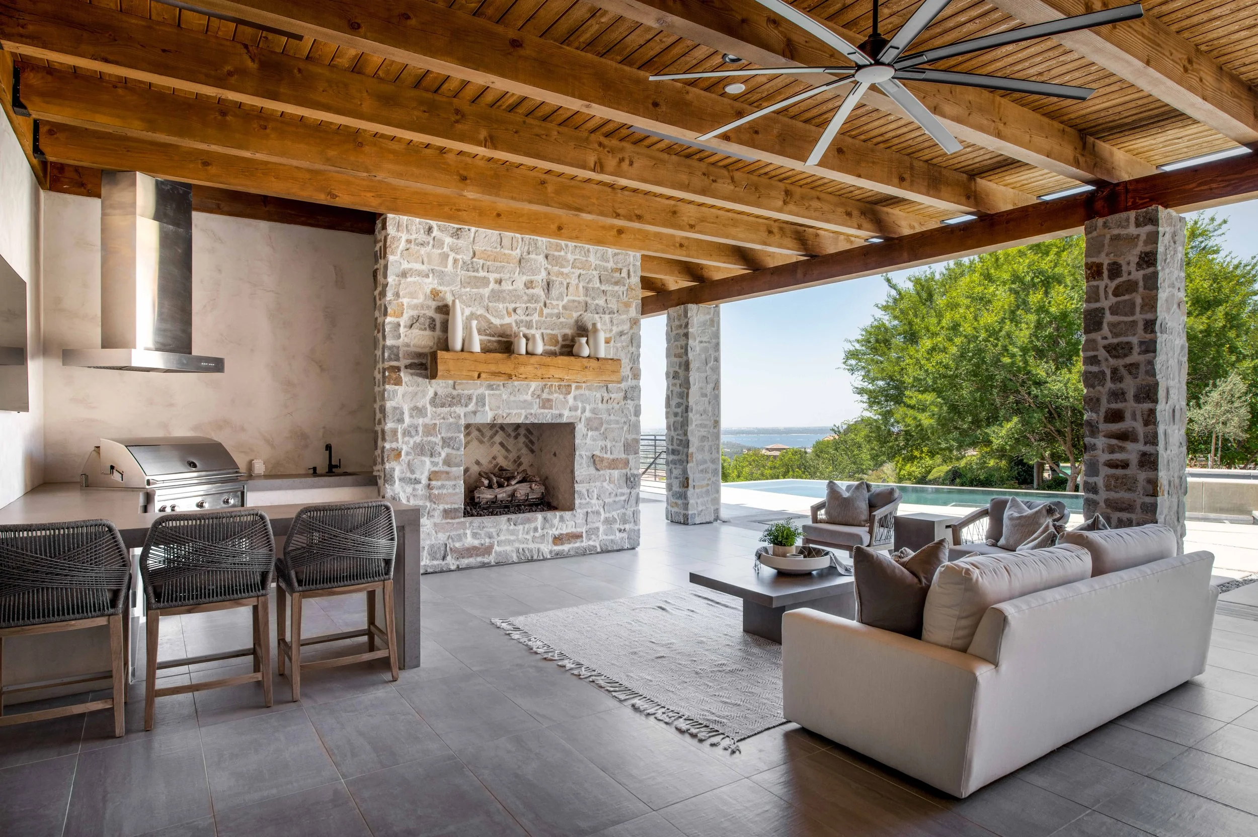 Open outdoor living space with a wooden ceiling, a ceiling fan, stone fireplace, lounge seating, an outdoor kitchen area, and trees in the background.
