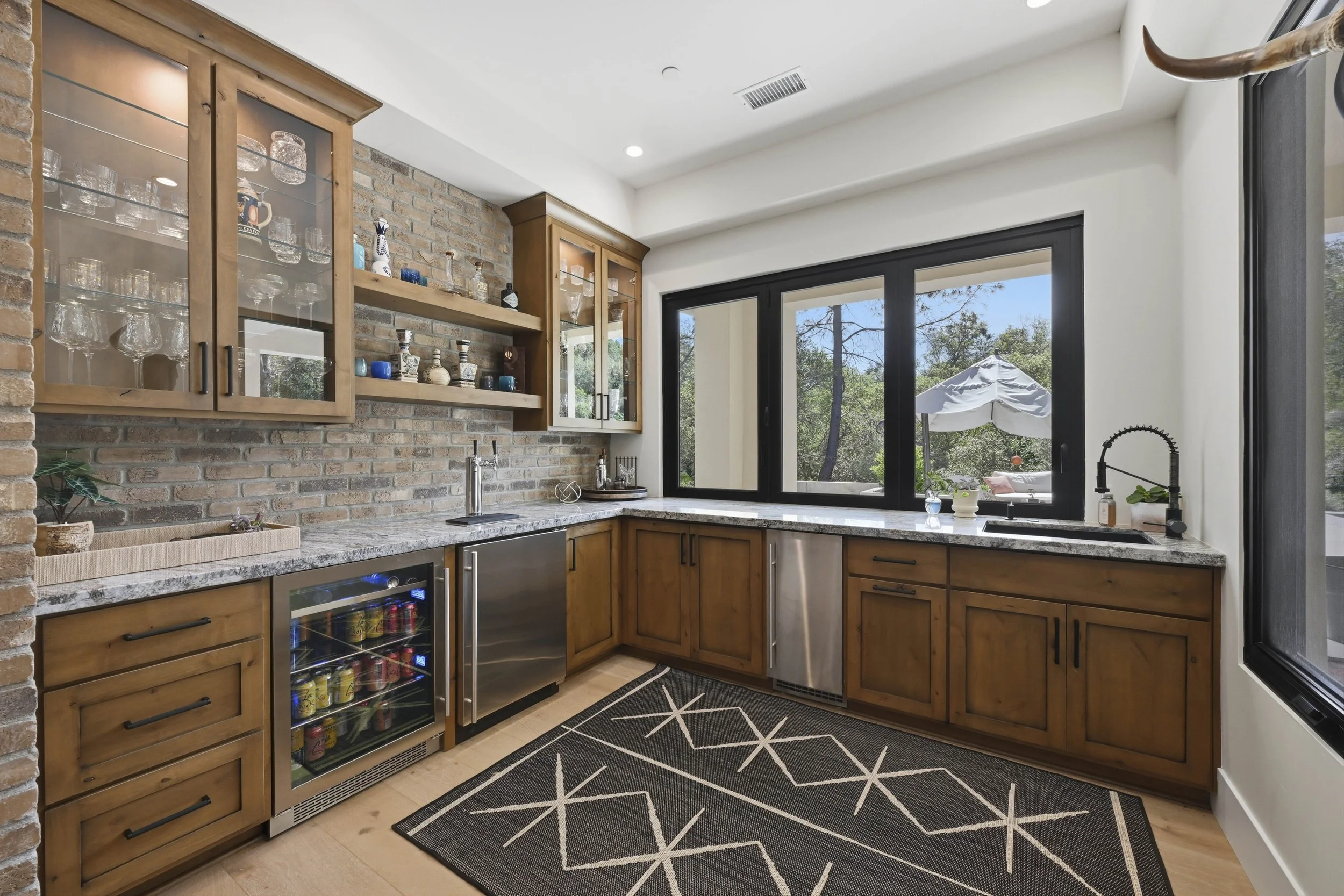 A modern kitchen with wooden cabinets, a brick wall, a marble countertop, a wine fridge, and large windows showing an outdoor patio with a white umbrella and trees.