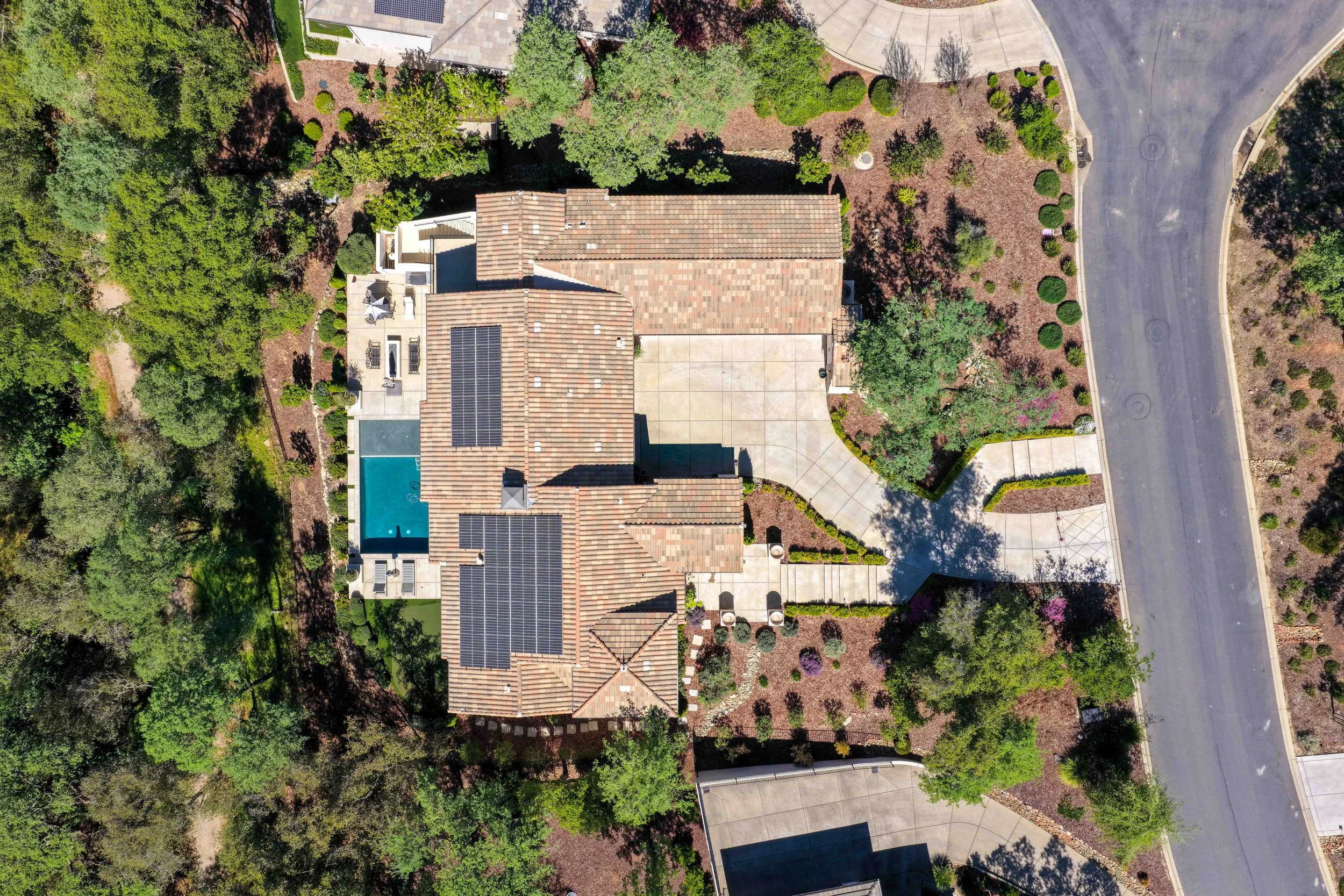 An aerial view of a residential property featuring a house with a tile roof equipped with solar panels, a backyard swimming pool, landscaped greenery, and surrounding trees, alongside a curving street.
