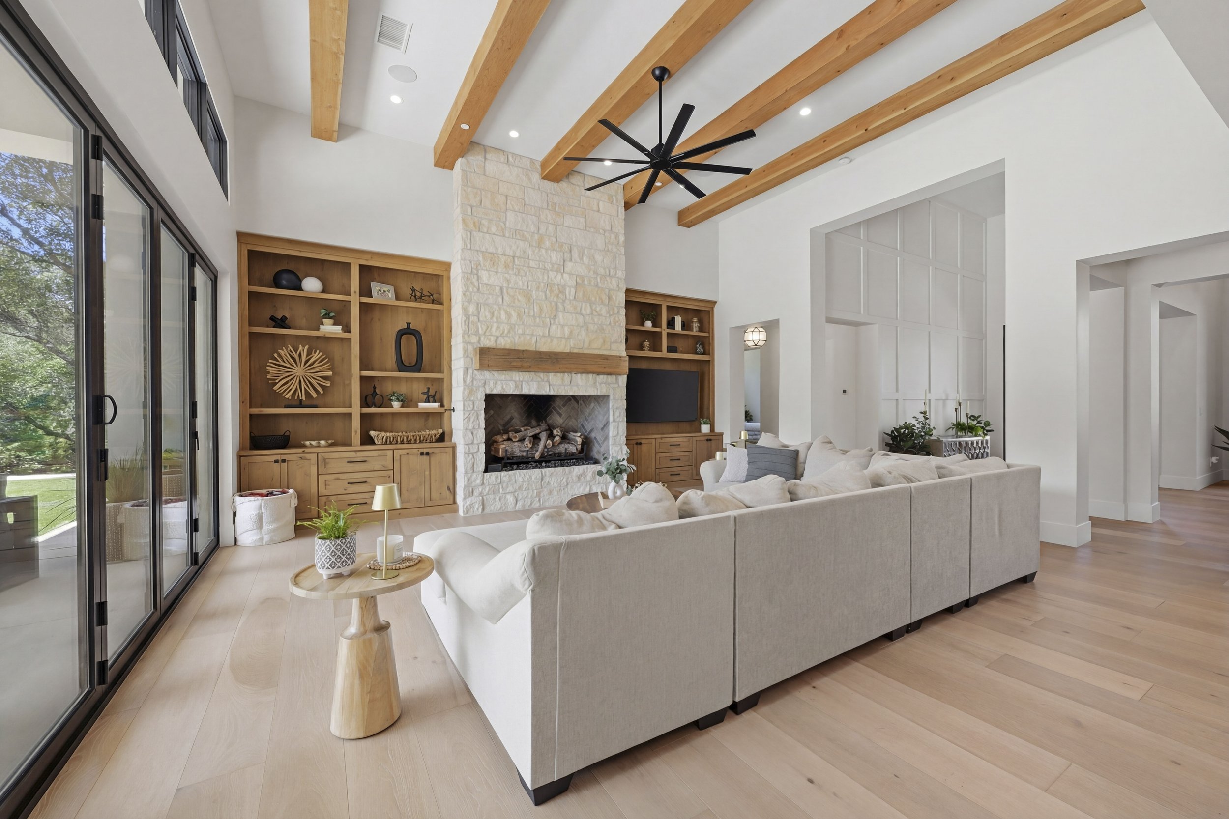 Living room with a vaulted ceiling, wooden beams, a ceiling fan, a stone fireplace, built-in wooden shelves, and sliding glass doors leading outside.