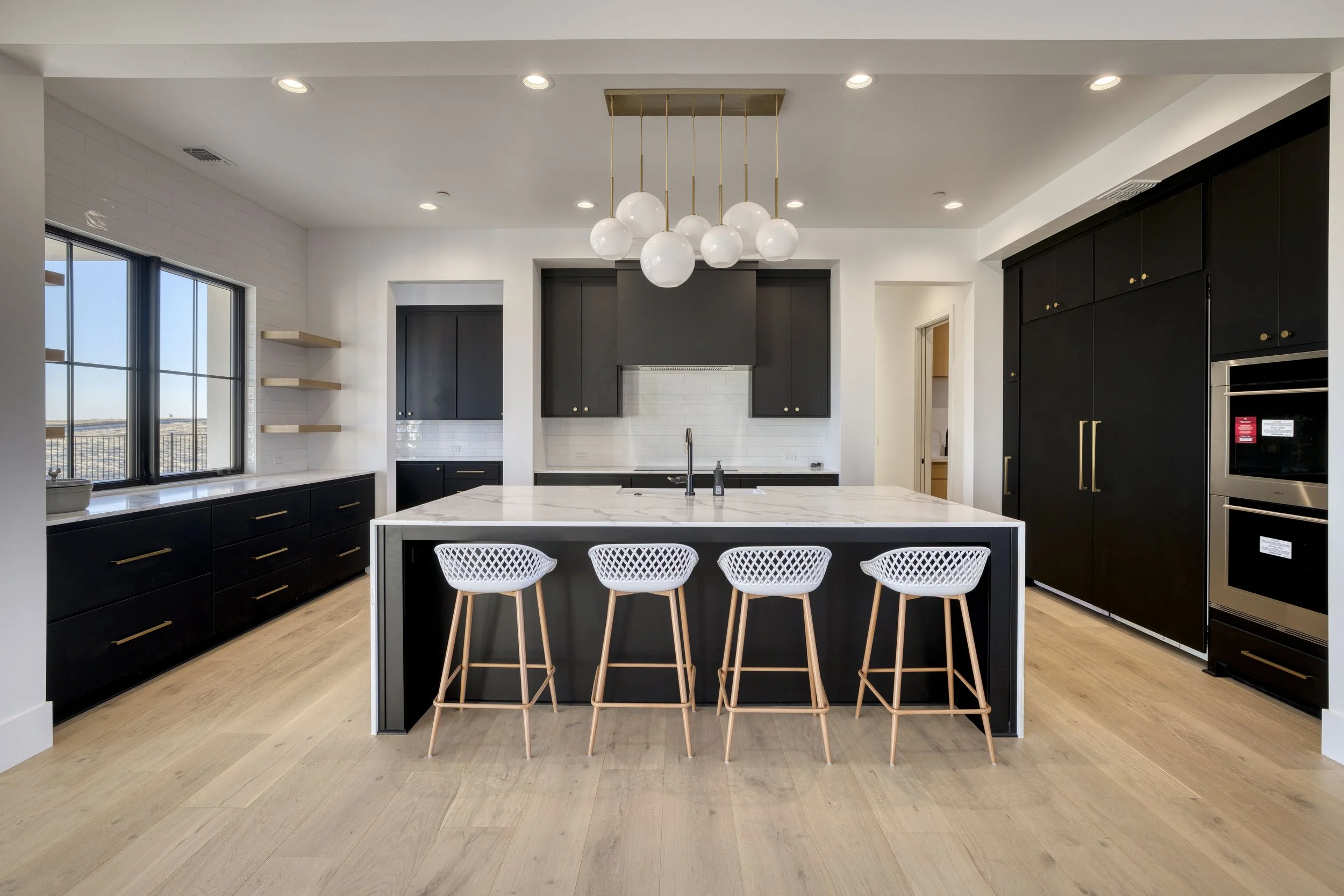 Modern kitchen with black cabinetry, white marble island, floating shelves, large window, and white chairs.