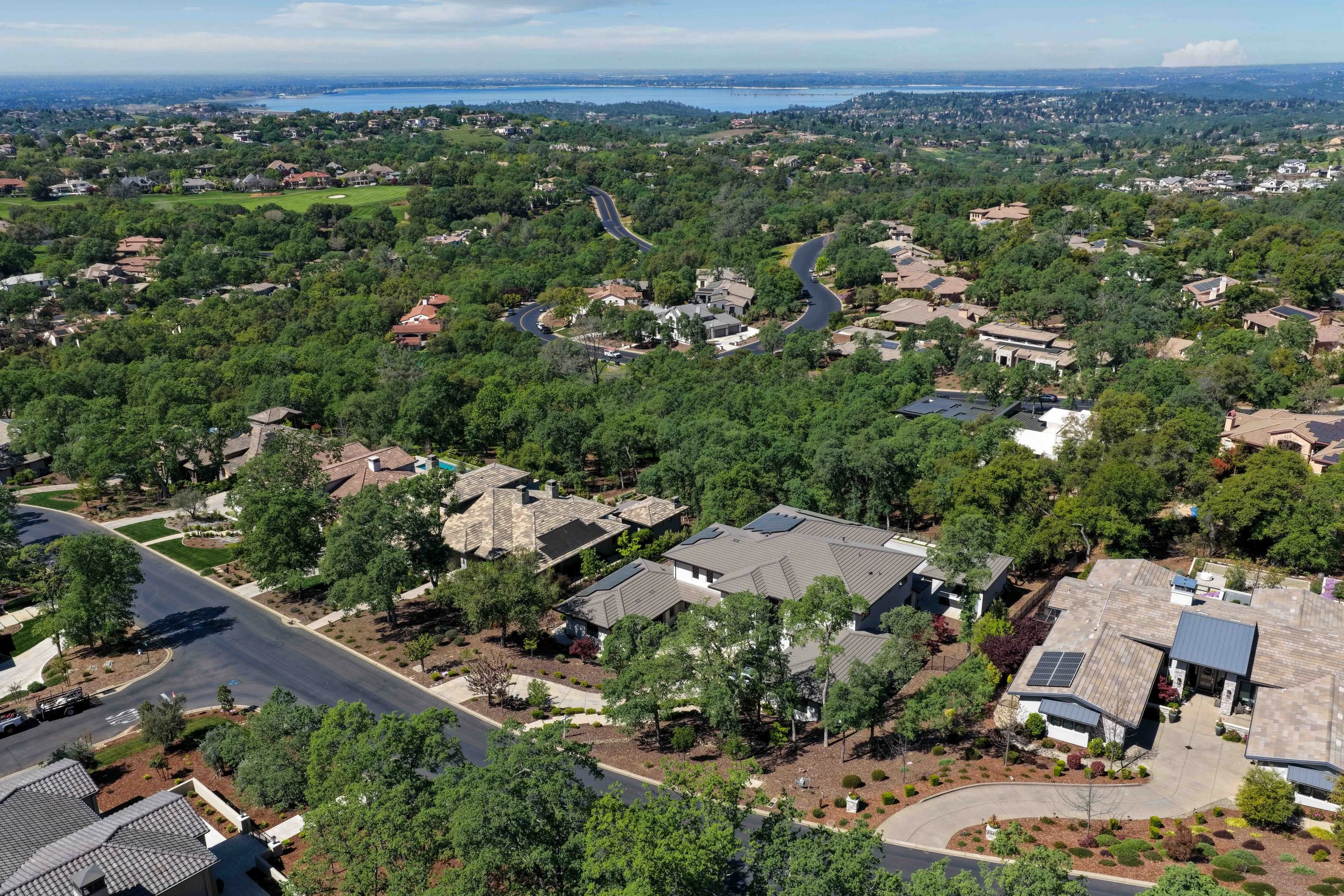 Aerial view of a suburban neighborhood with winding roads, numerous houses surrounded by trees, green spaces, and a distant body of water under a partly cloudy sky.