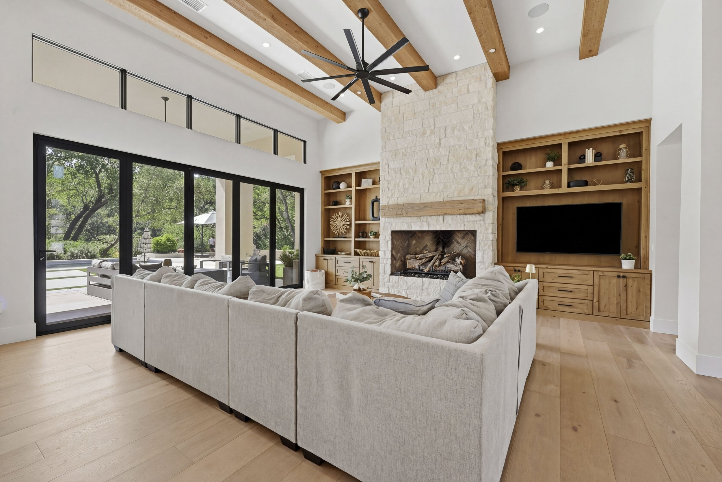 Living room with large white sectional sofa, a fireplace with a wood mantel, and built-in wooden shelves with decorative items. Large sliding glass doors open to an outdoor patio with trees and outdoor furniture.