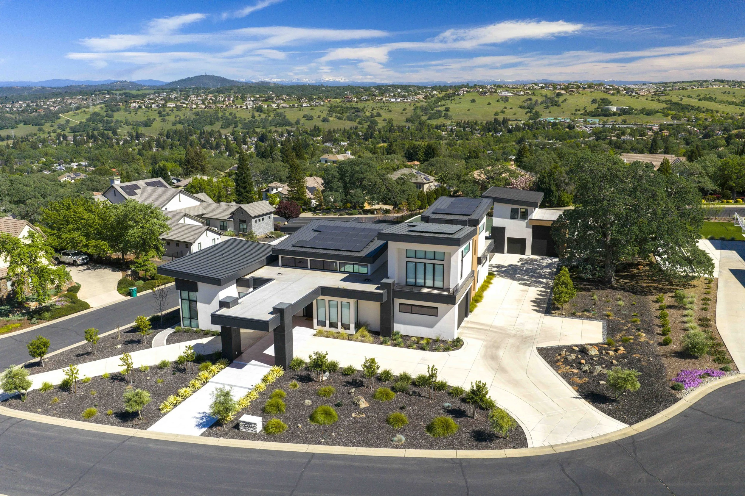 Aerial view of a modern house with solar panels, surrounded by landscaped gardens, in a suburban neighborhood with green hills and houses in the background under a blue sky.