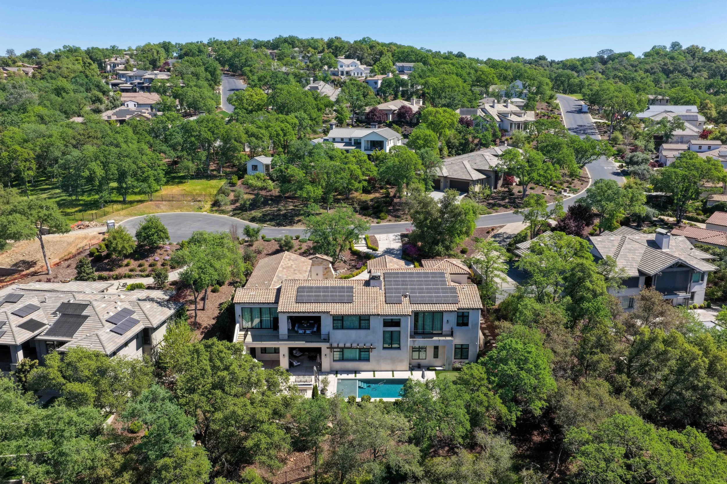 An aerial view of a suburban neighborhood with numerous houses surrounded by lush green trees, multiple winding streets, and a clear blue sky.