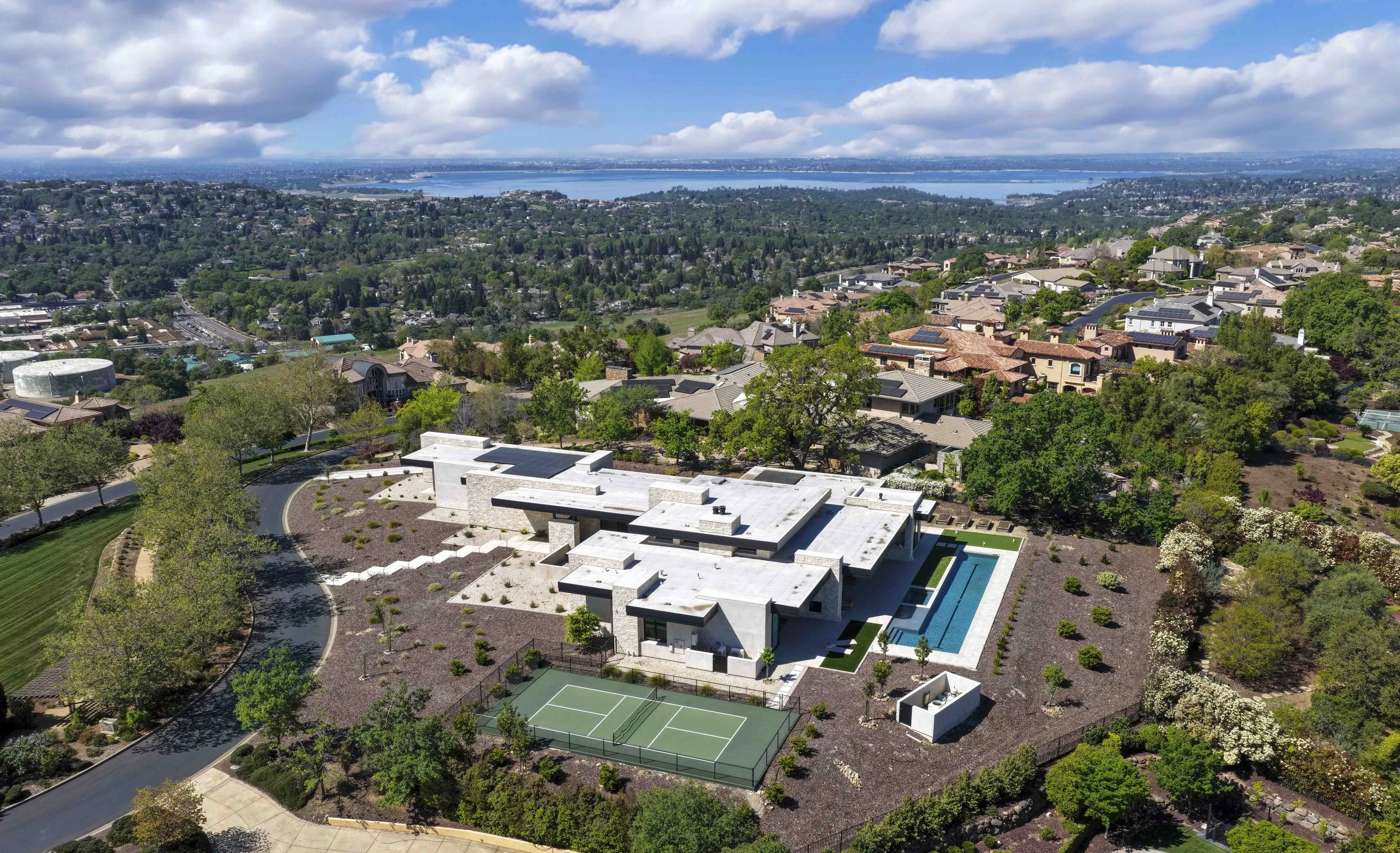 Aerial view of a residential neighborhood with a modern white house, a swimming pool, tennis court, green trees, and a scenic body of water in the background under partly cloudy skies.