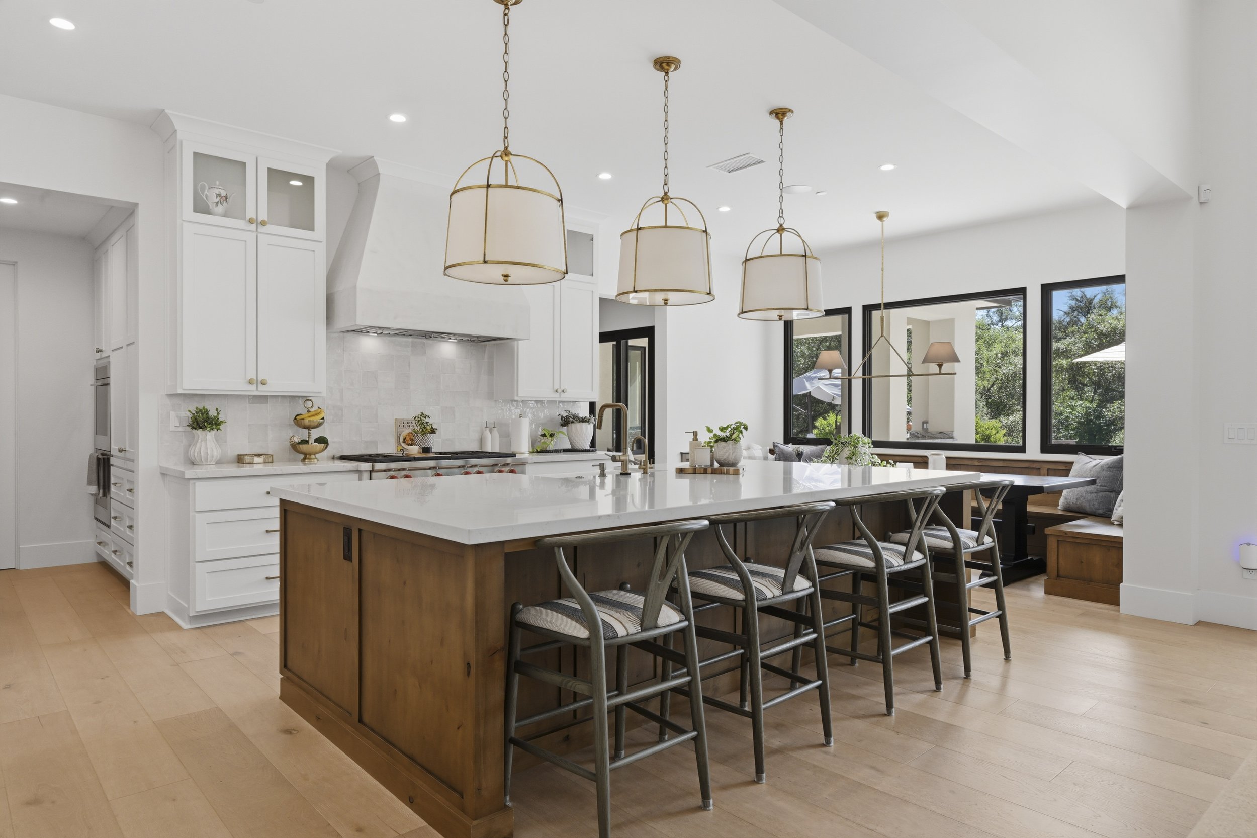 Modern open-concept kitchen and dining area with white walls, large windows, and wooden flooring. The kitchen features a central island with a wooden base, white countertop, and barstool seating. Hanging above are three stylish pendant lights with wh