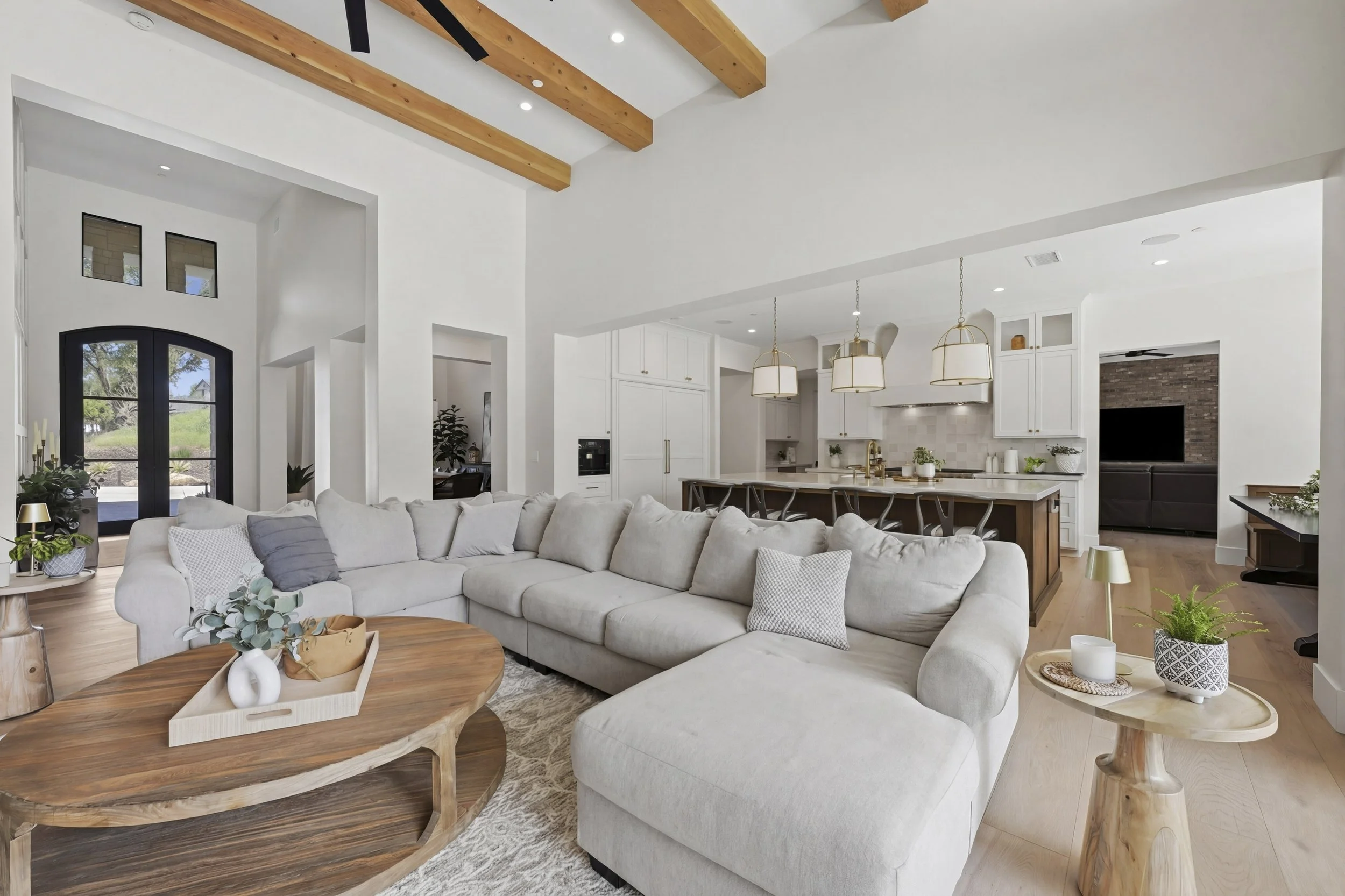 Bright, open-concept living room and kitchen with white walls, high ceilings with wooden beams, large window, and modern furniture.