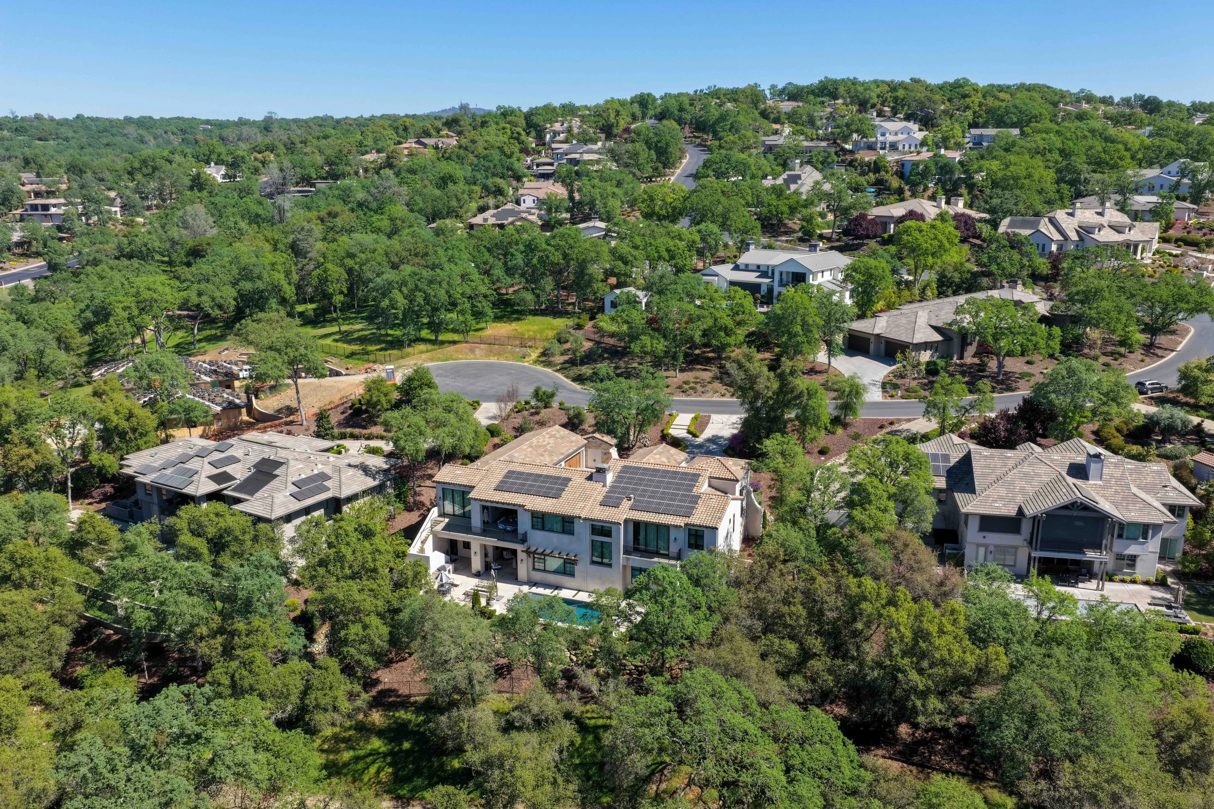 Aerial view of a suburban neighborhood with numerous houses surrounded by green trees and landscaping, under a clear blue sky.