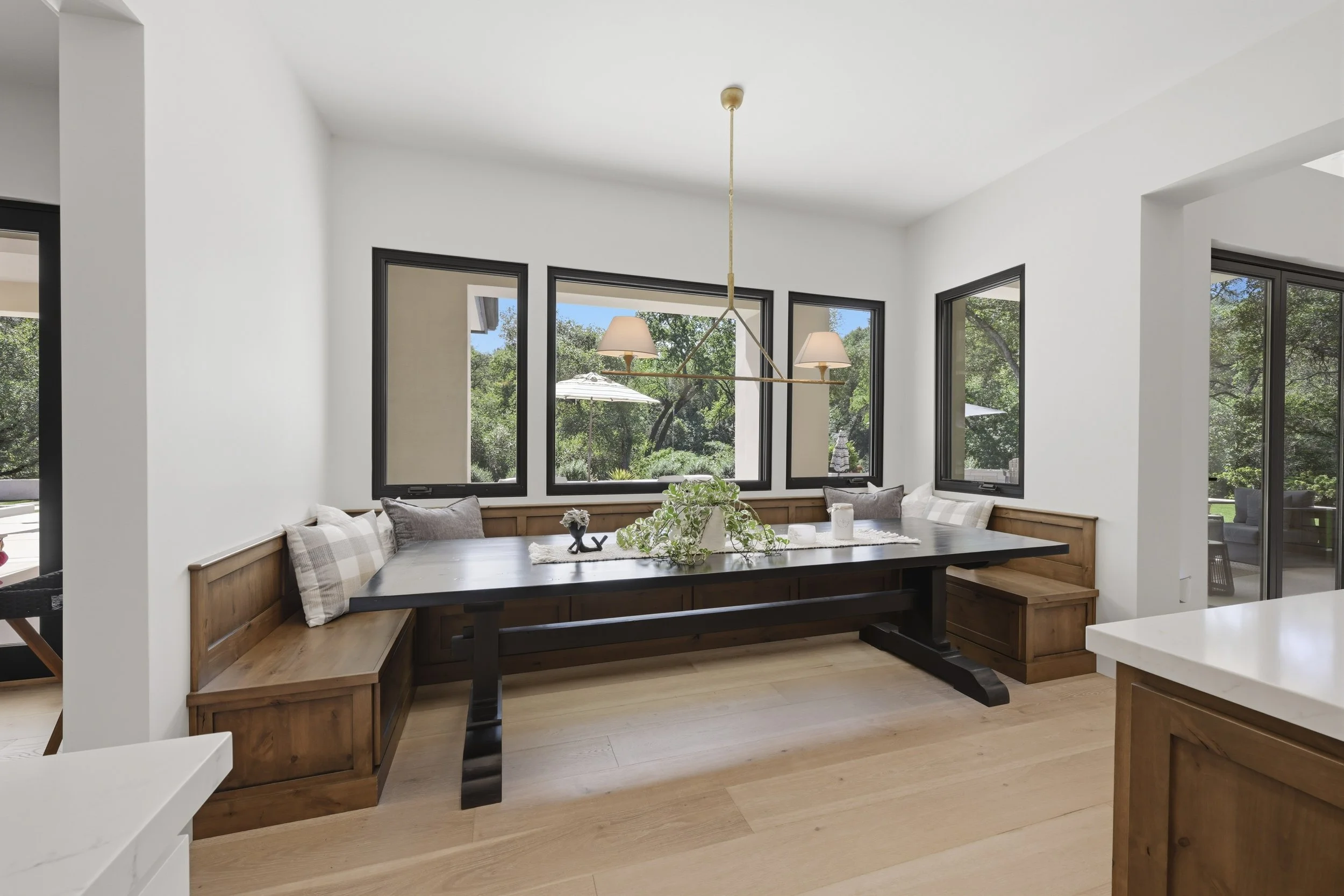 Bright dining area with a wooden bench and table, decorated with cushions, plants, and candles, surrounded by large windows showing green trees outside.