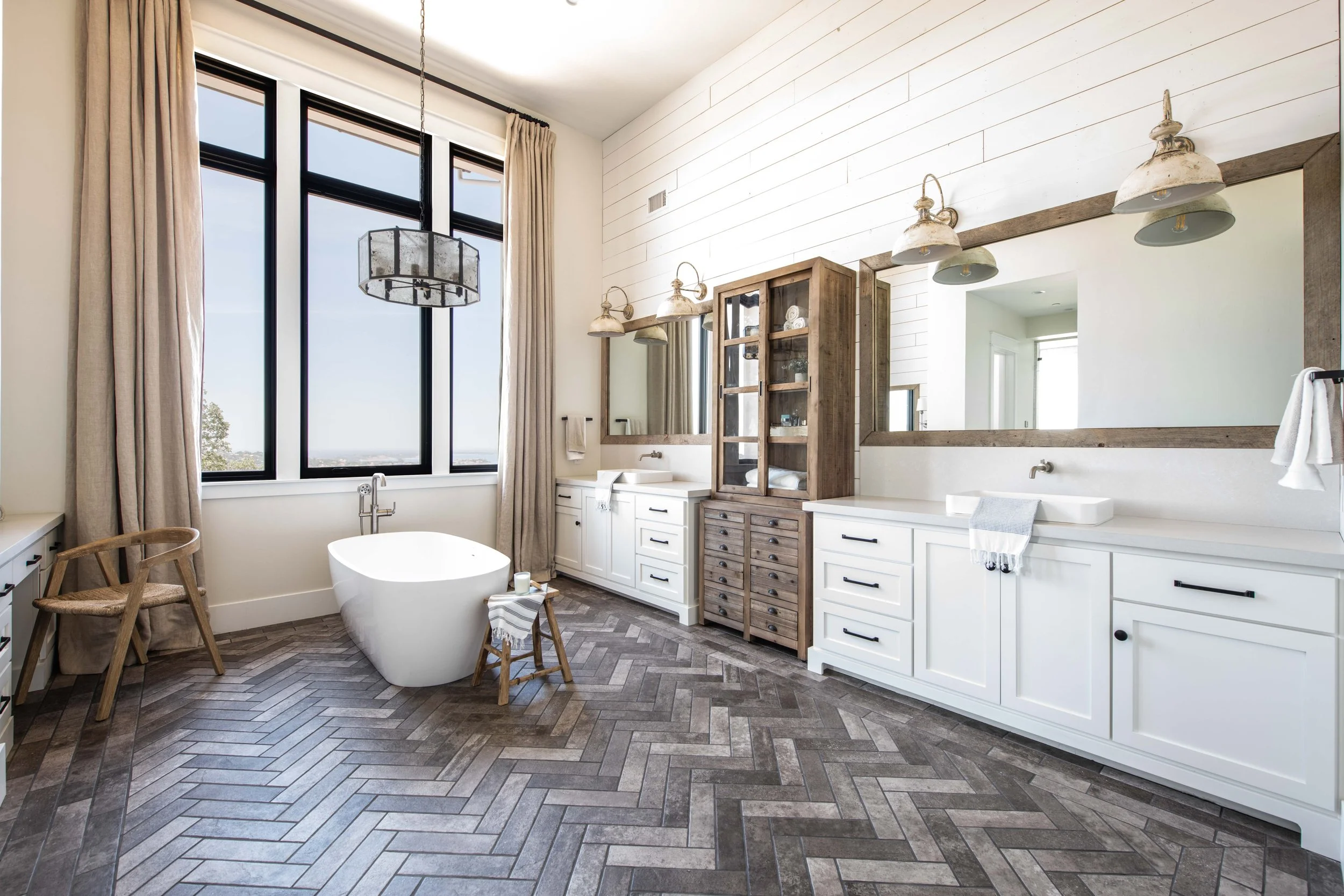 A spacious bathroom with large windows, a freestanding bathtub, wooden and white cabinetry, and a large mirror, with earthy tones and modern fixtures.