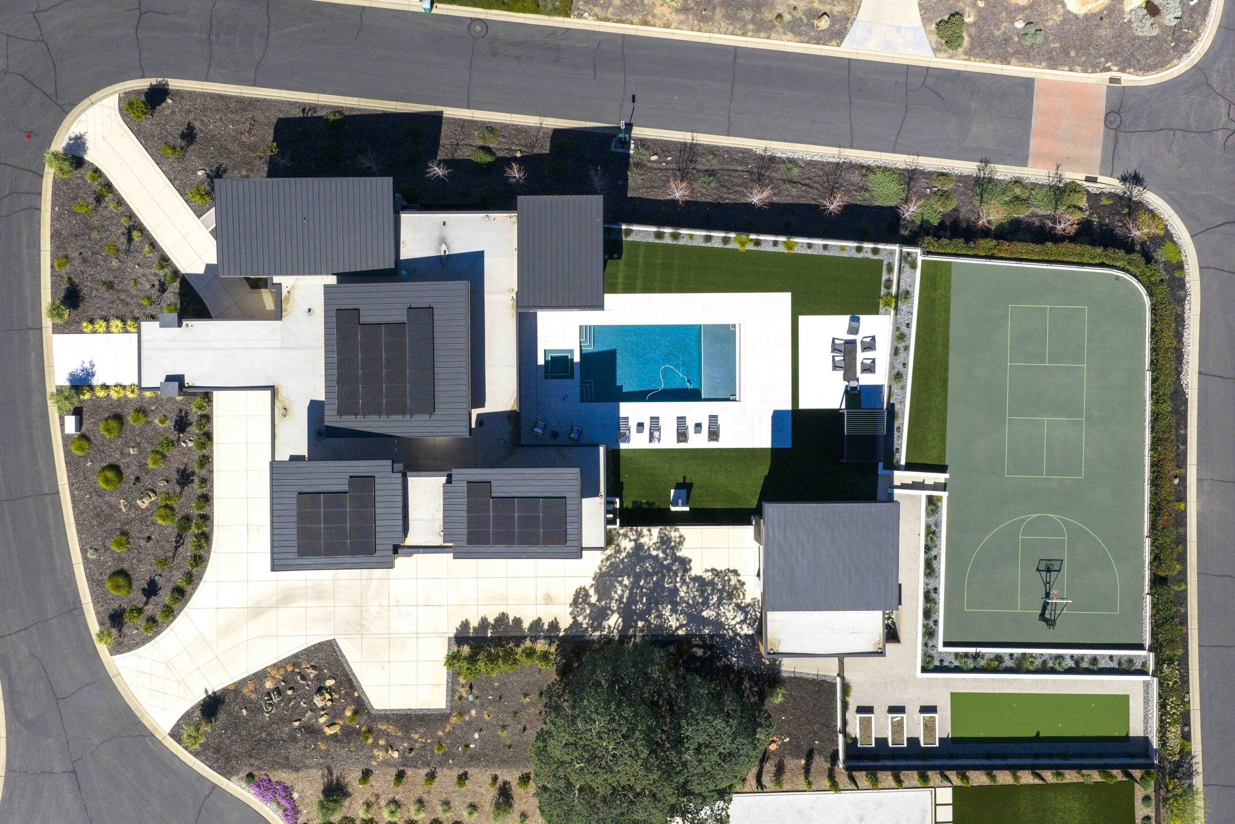 Aerial view of a modern residential property with a swimming pool, basketball court, landscaped garden, and surrounding paved pathways and roads.