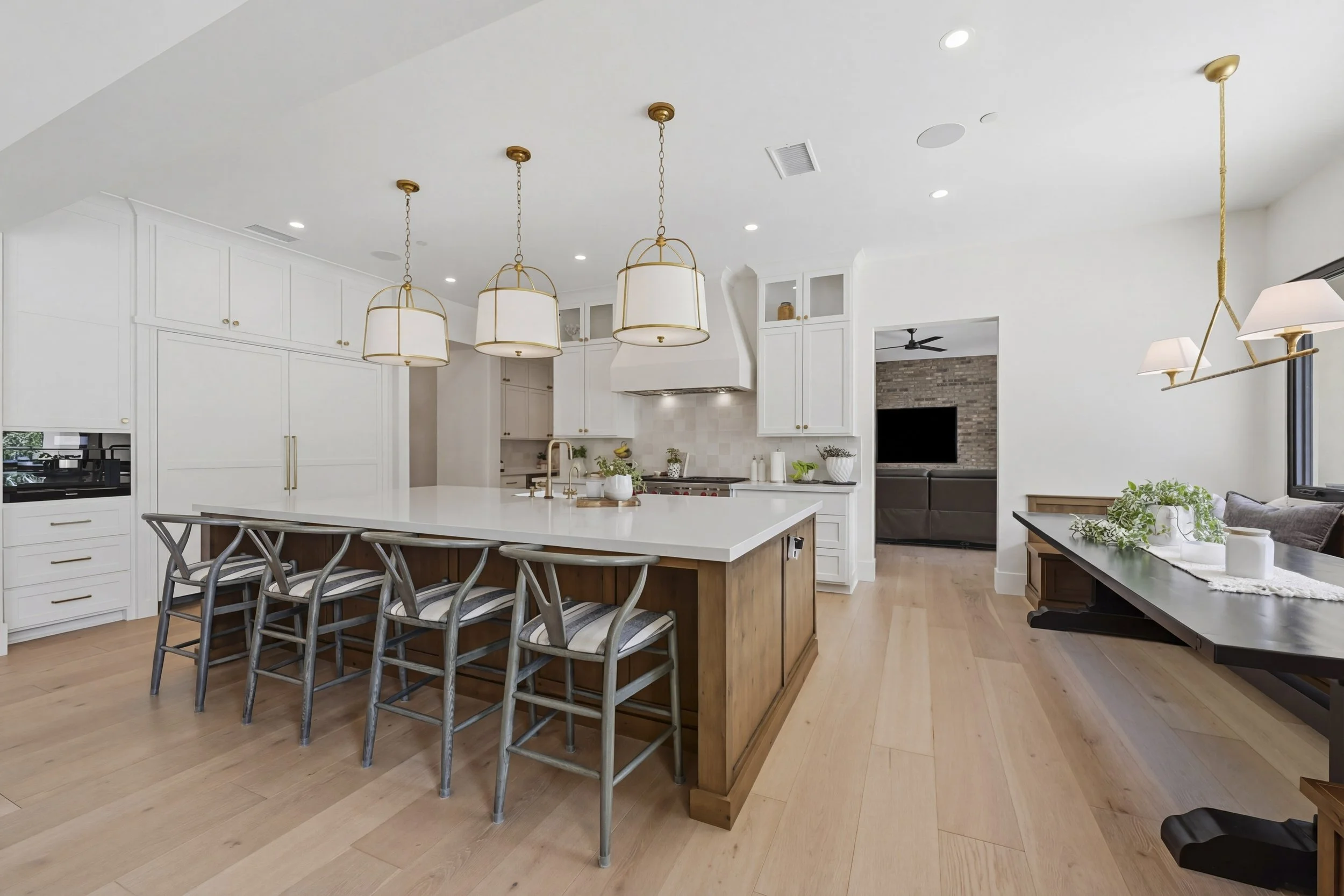 Modern open-plan kitchen with white cabinetry, a large island with seating, three gold and white pendant lights, and a seating area with a TV and brick wall in the background.