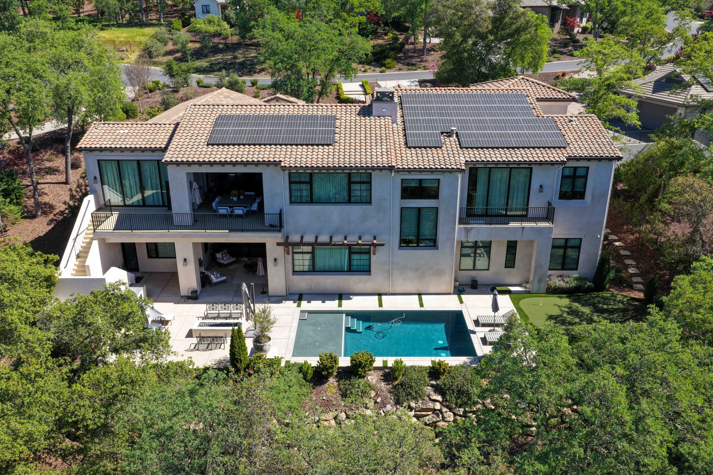 Aerial view of a modern house with a tiled roof and solar panels, surrounded by trees, a backyard with a swimming pool, patio, and outdoor furniture.
