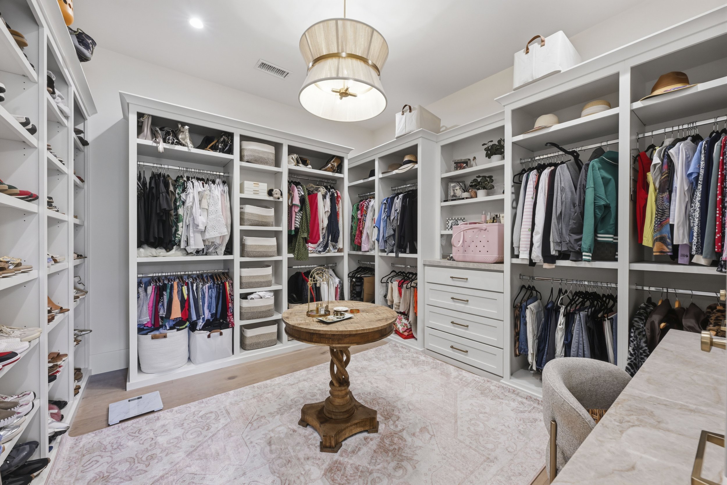 A walk-in closet with white shelving units filled with clothes, shoes, hats, and storage baskets. There is a round wooden table in the center, a soft beige rug on the floor, and a light fixture hanging from the ceiling.