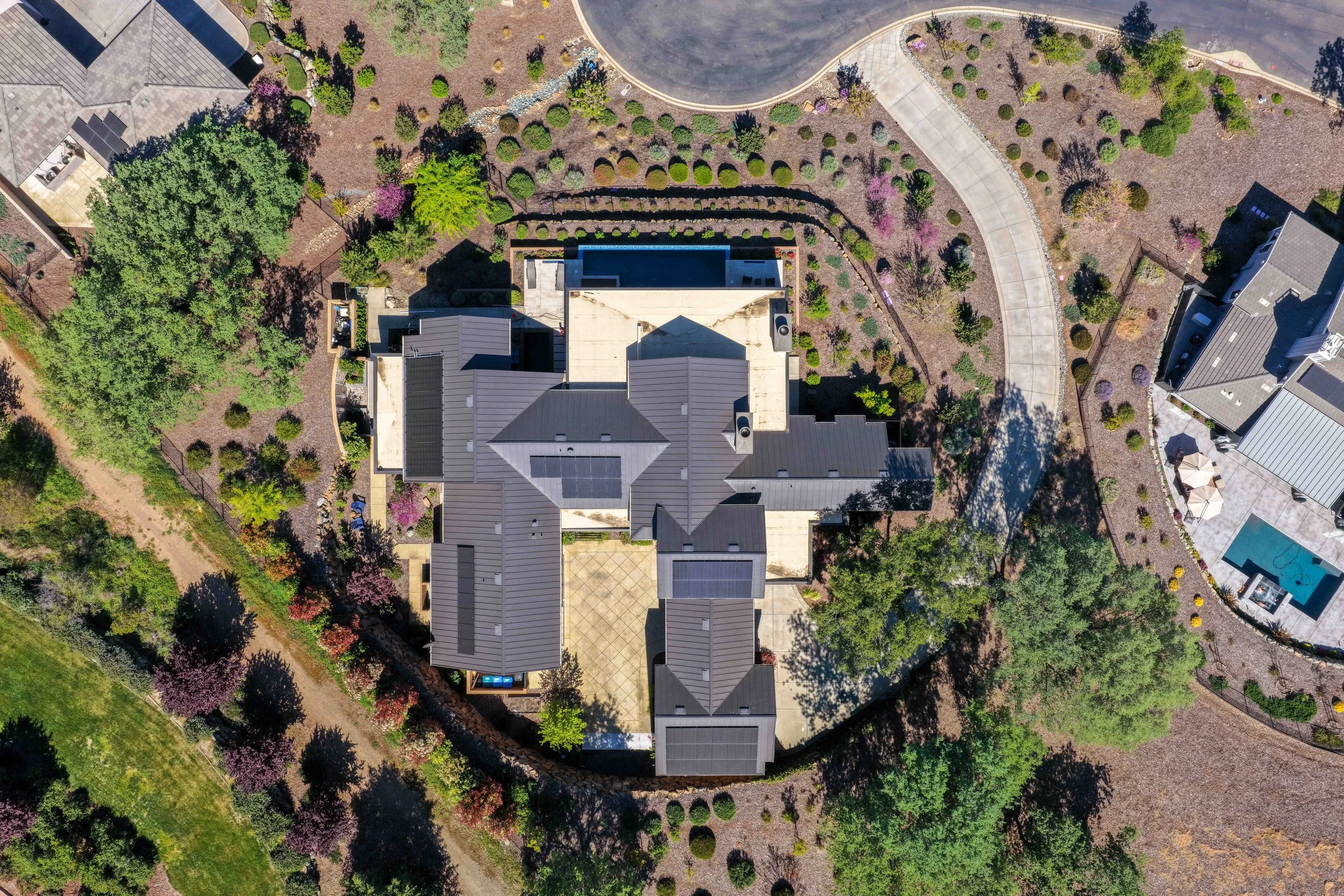 An aerial view of a residential backyard with trees, shrubs, flower beds, a swimming pool, and a house with solar panels on the roof.