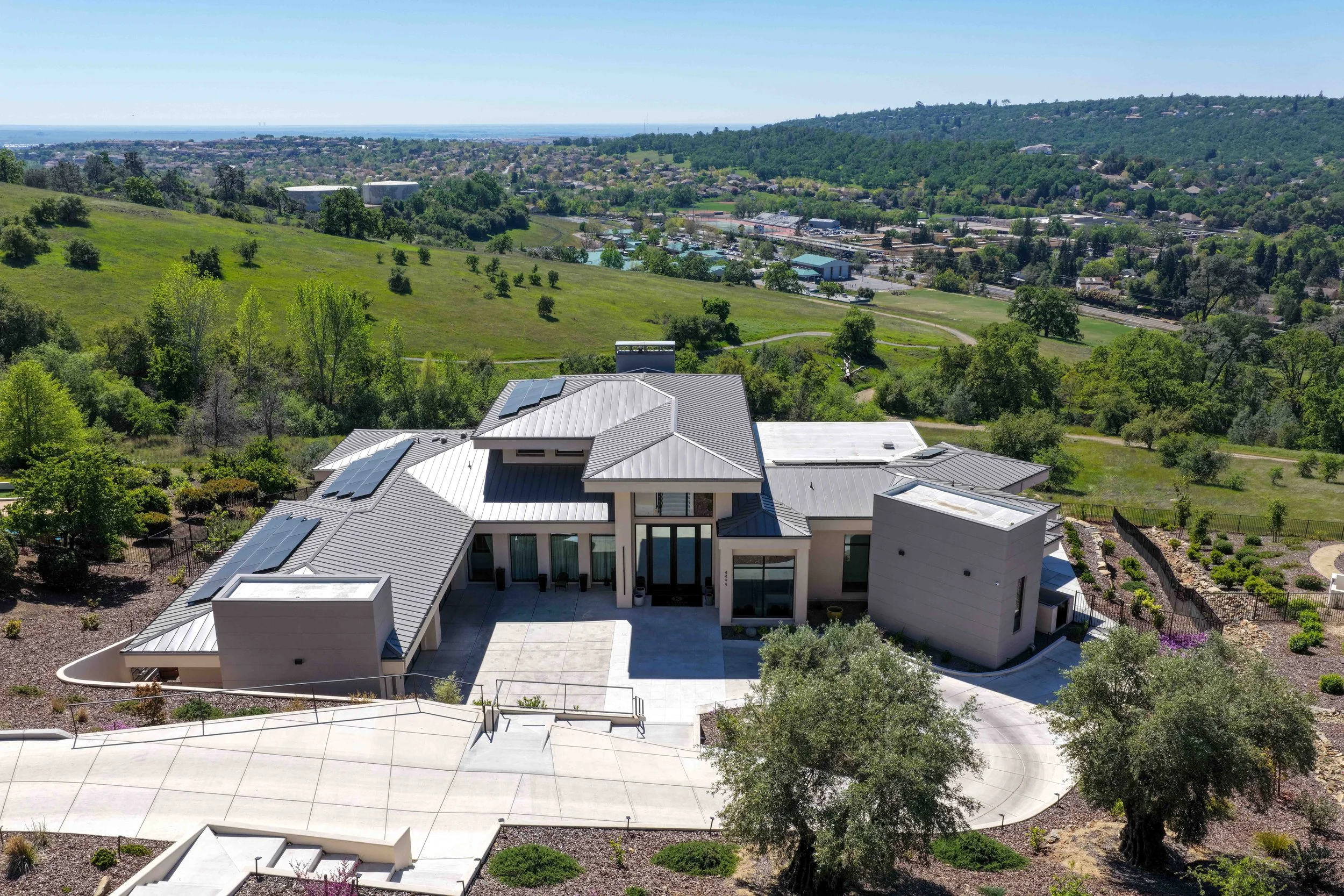 Aerial view of a modern house with solar panels on the roof, surrounded by a landscaped yard with trees and shrubs, overlooking a green hillside, a neighborhood, and distant hills and ocean in the background.