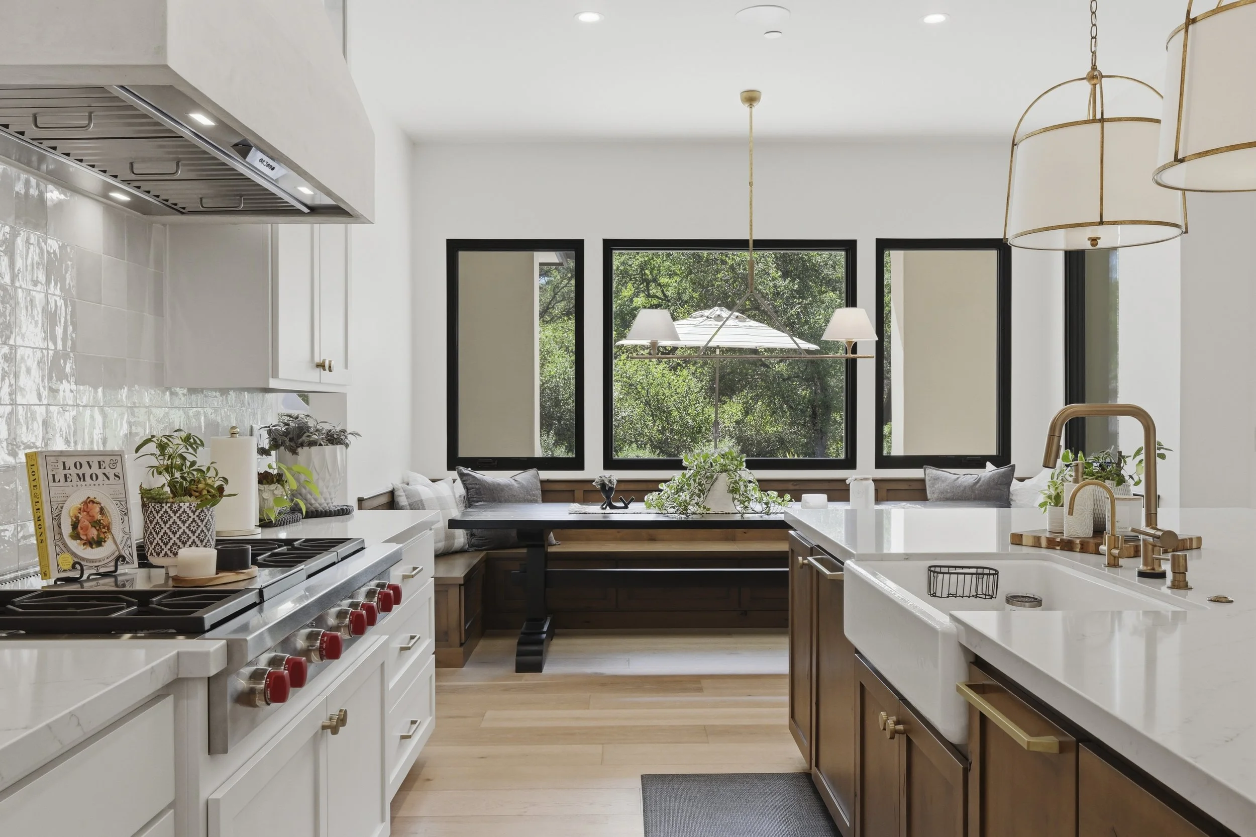 Modern kitchen with white cabinets, a marble countertop, wooden accents, and large windows overlooking greenery. There are pendant lights hanging from the ceiling.