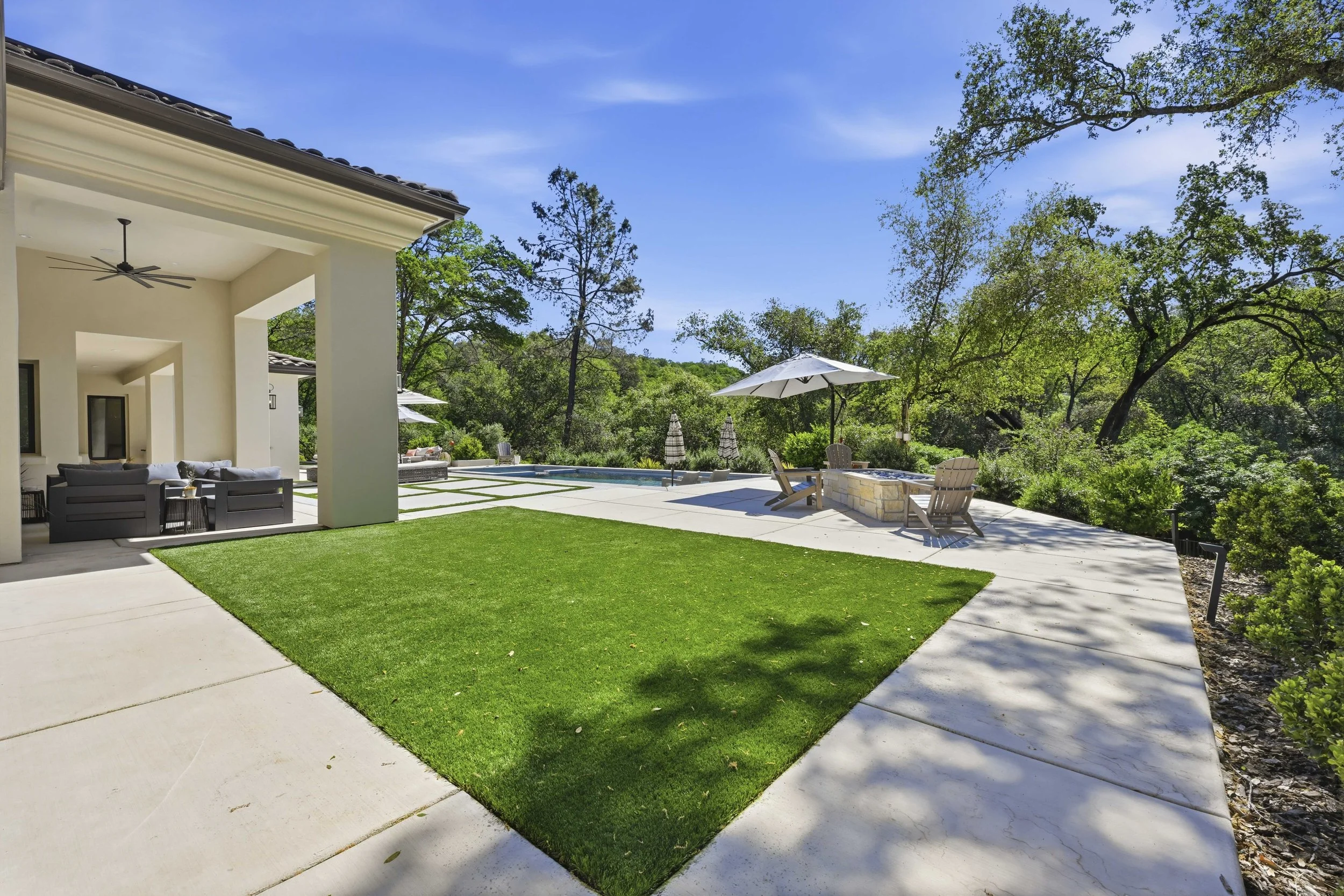 A backyard patio area with a covered outdoor seating area, a swimming pool, patio chairs with umbrellas, and surrounded by trees under a blue sky.