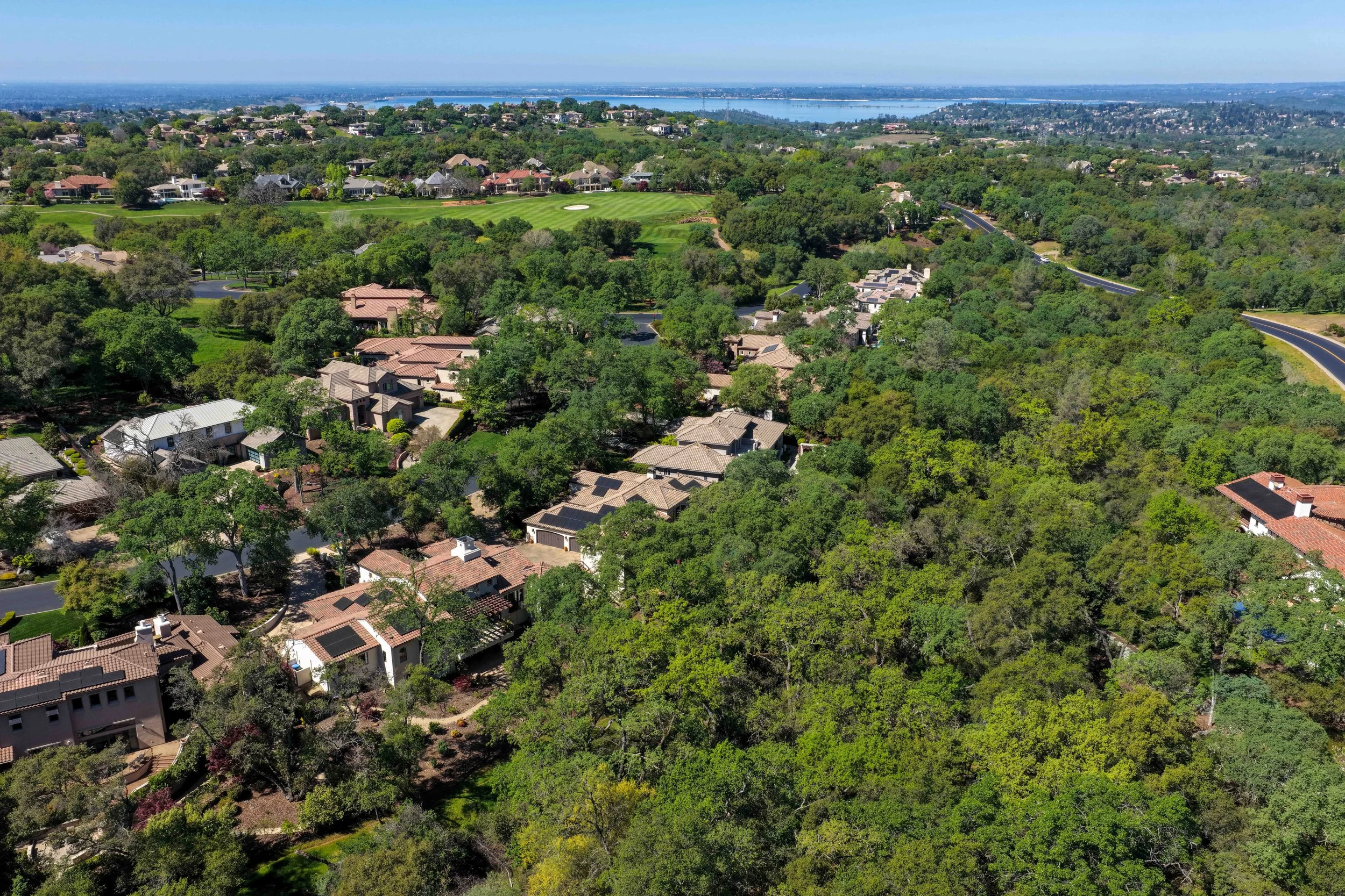 Aerial view of a suburban neighborhood with many houses surrounded by green trees, a golf course, and a body of water in the distant background on a clear, sunny day.