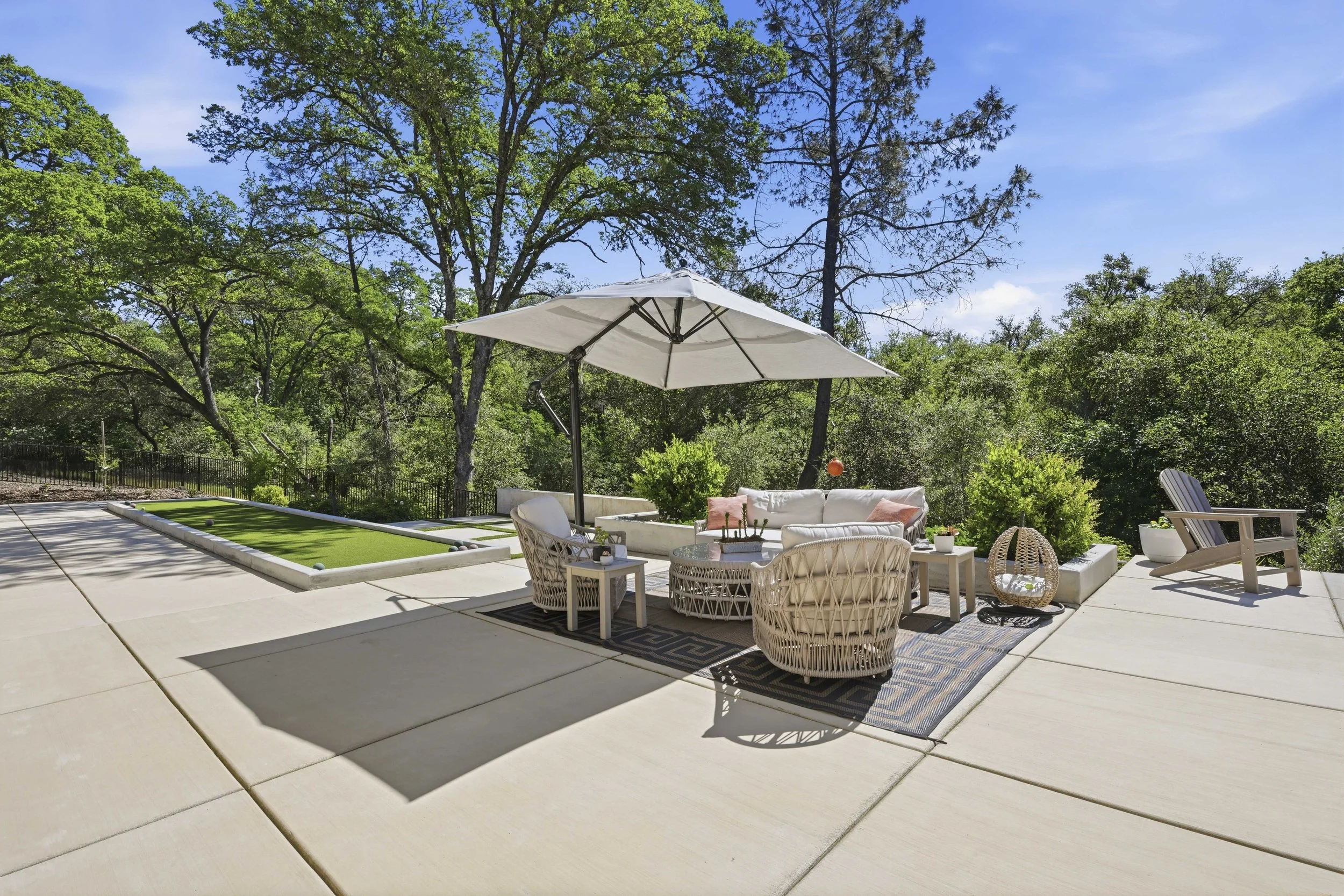 An outdoor patio with white furniture, including a sofa, chairs, and a table with a black and white rug underneath. A white patio umbrella provides shade. There is a small putting green with golf balls on the left side. Surrounding the patio are tall