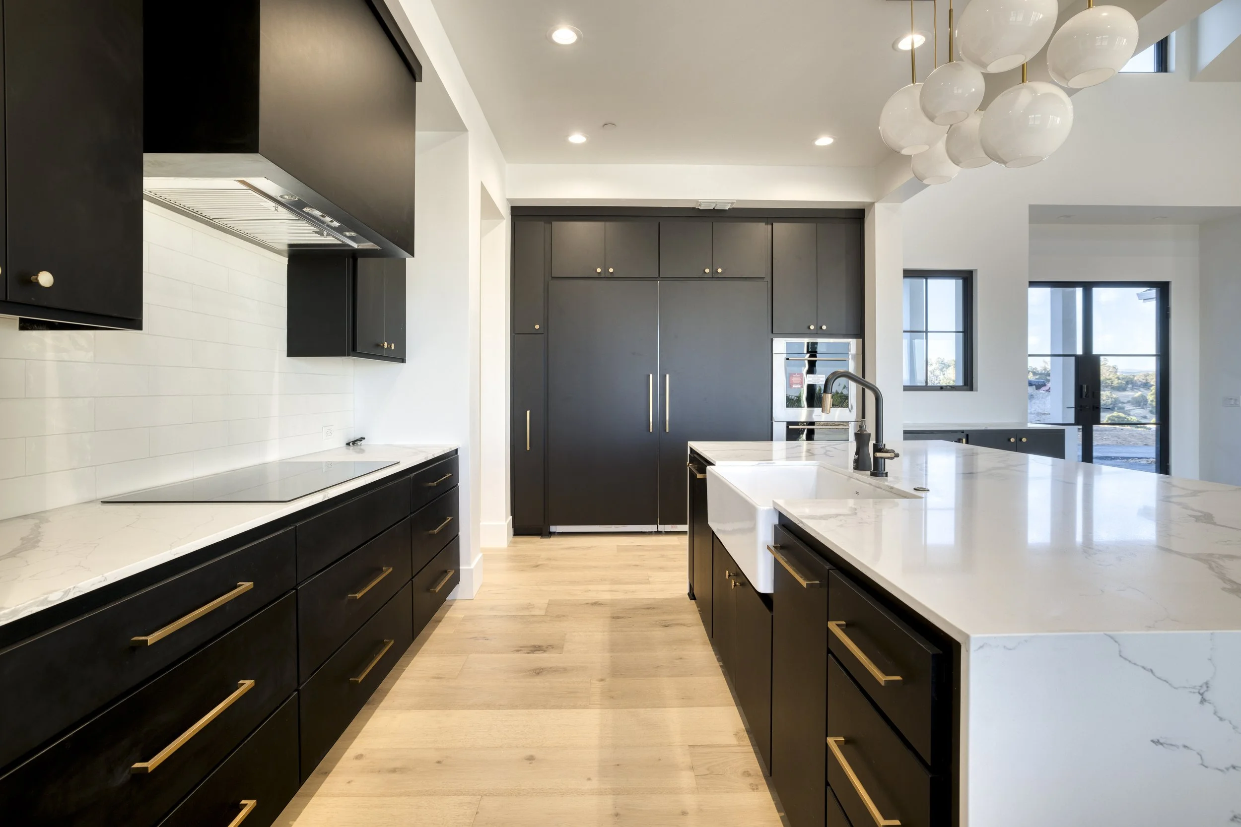 Modern kitchen with black cabinets, white marble countertops, a large integrated refrigerator, a built-in oven, and a white sink with a black faucet. There are large windows with a view outside, and a contemporary light fixture with white globes hang