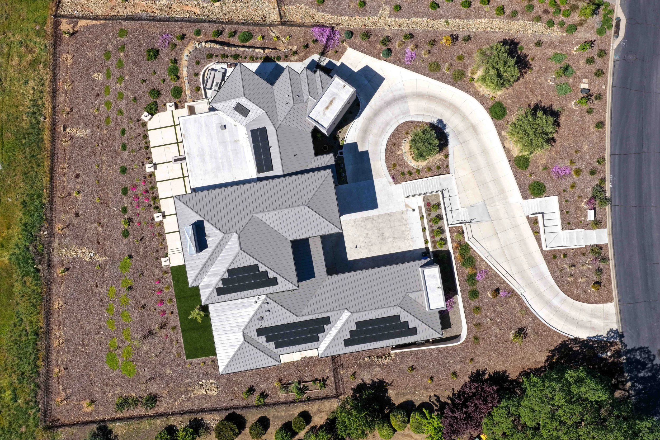 Aerial view of a large house with a gray roof, surrounded by landscaped yard with small trees, bushes, purple flowering plants, concrete pathways, and a street on the right side.