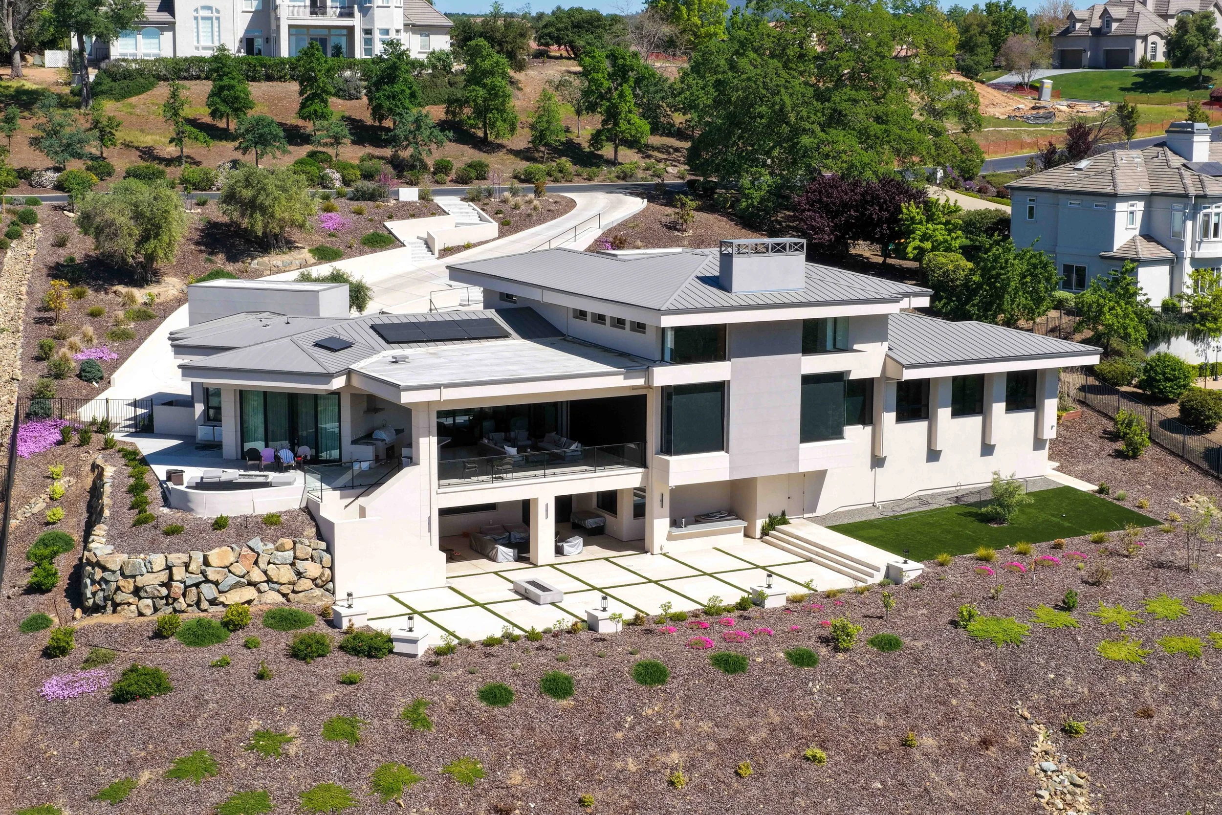 A modern, multi-level house with a gray metal roof and large windows, surrounded by landscaped gardens, trees, and a patio with outdoor furniture.