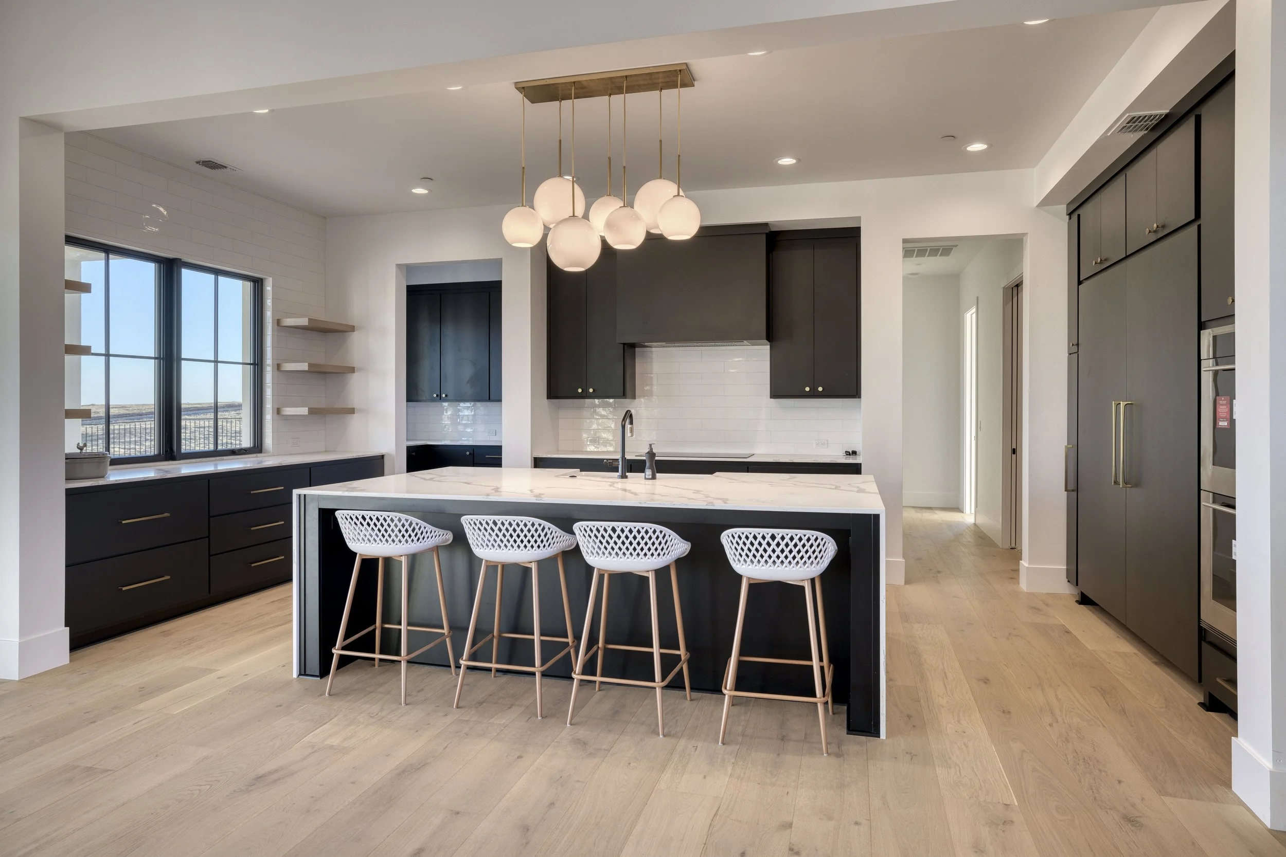 Modern kitchen with black cabinets, white island with marble countertop, four white woven bar stools with wooden legs, large window with black frame, light wood flooring, and a contemporary chandelier with white globes hanging from a wooden fixture.
