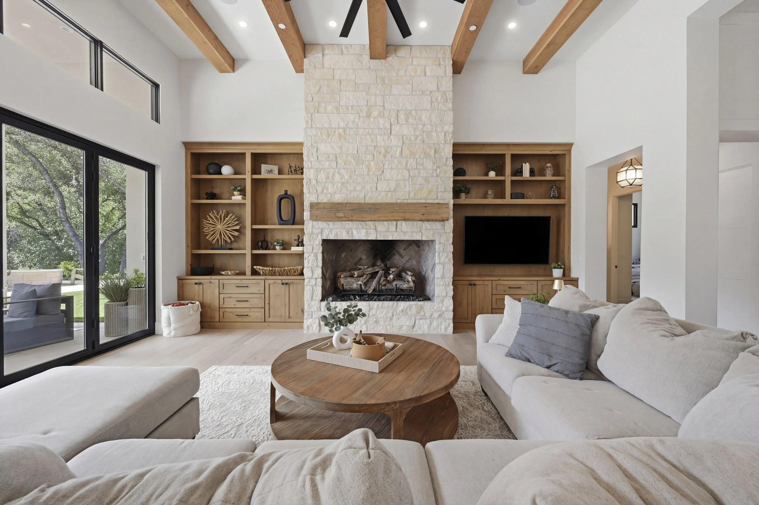 Modern living room with a white sectional sofa, a round wooden coffee table, a fireplace with a stone surround, and built-in wooden shelves. Large sliding glass door opens to an outdoor patio with trees visible outside.