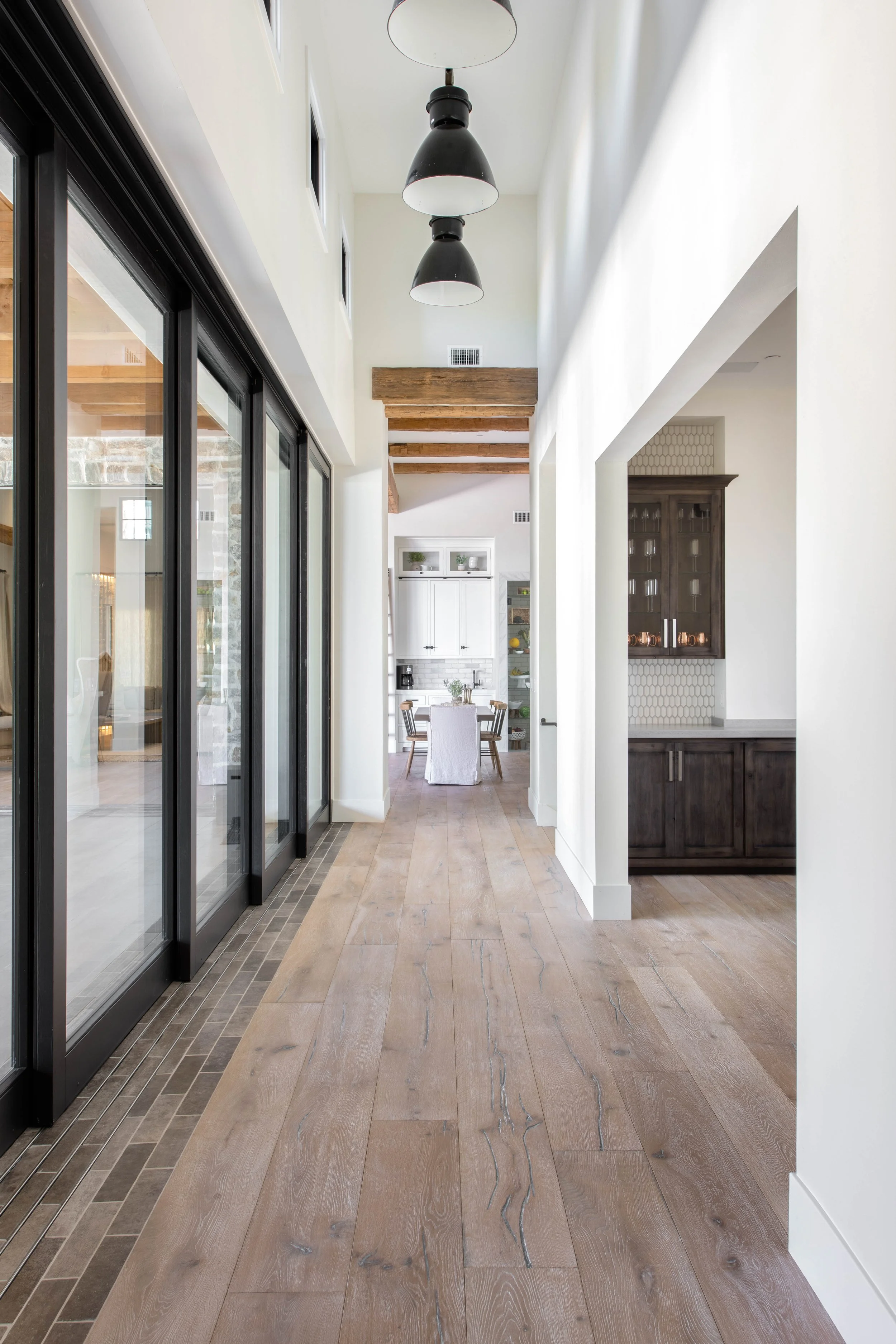 A hallway with light wood flooring, black-framed glass doors on the left, and white walls. There are wooden beams on the ceiling. In the background, a dining area with a white table and chairs is visible.