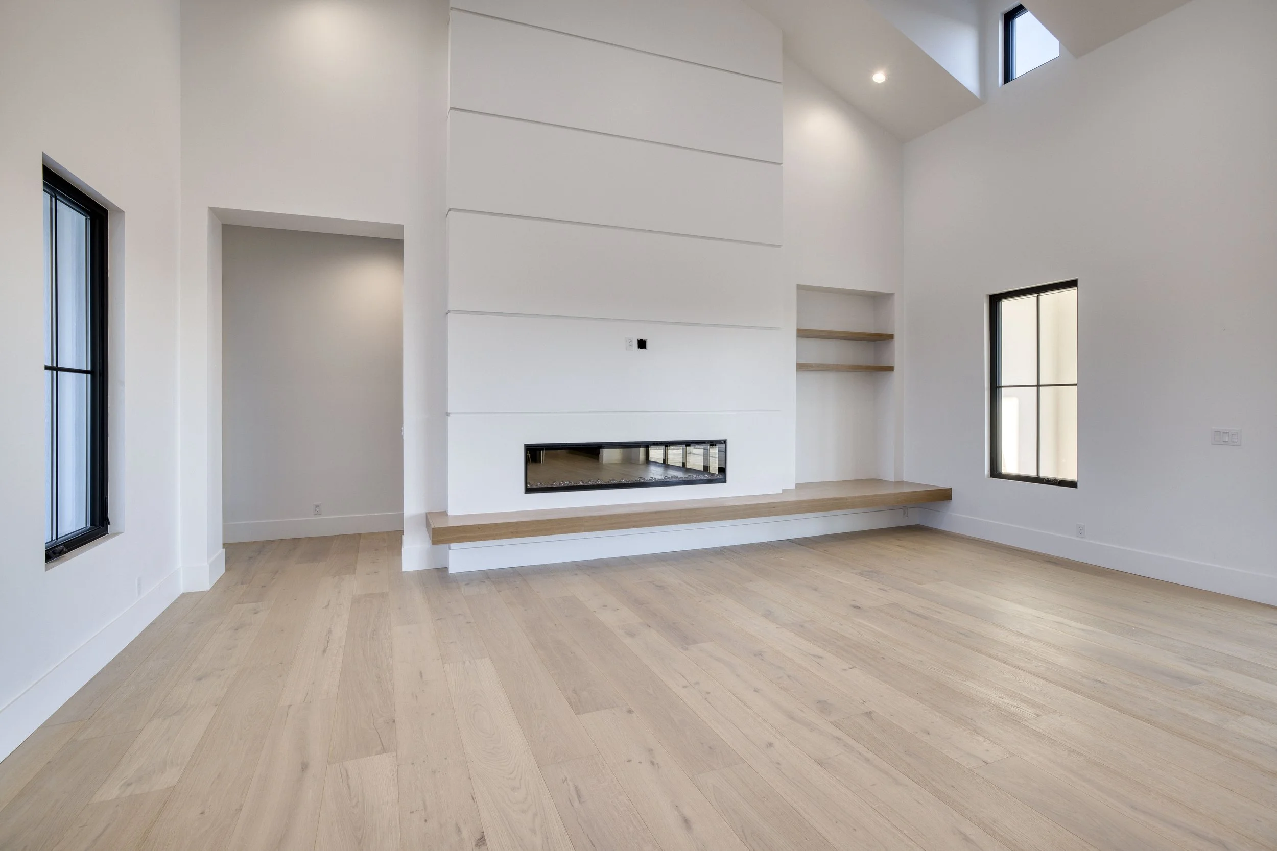 Interior of a modern, minimalistic room with white walls and light wooden flooring, large windows, and a built-in fireplace.