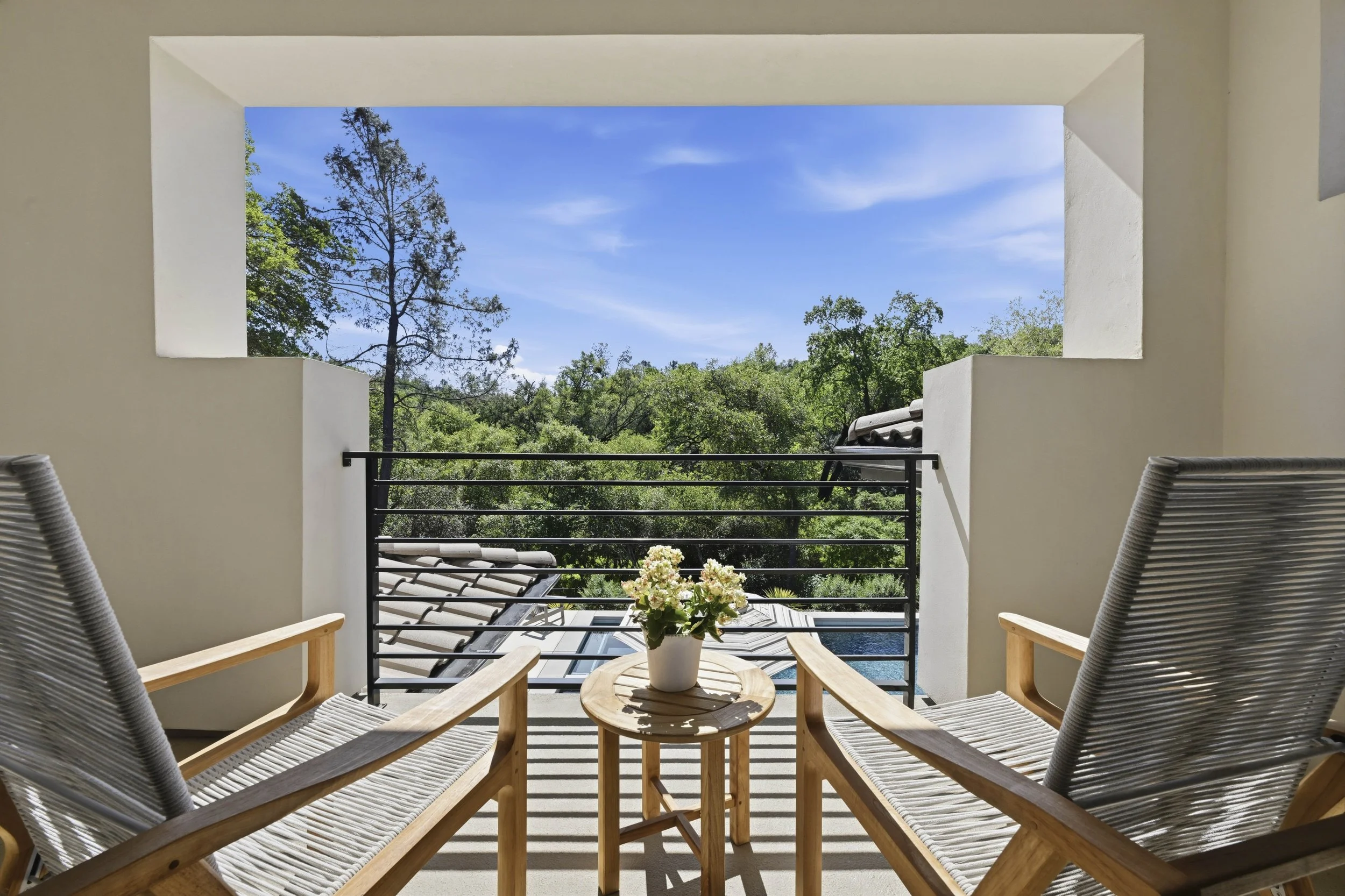 Balcony with two wooden chairs with woven seats facing outward, a small wooden round table with a potted white flower, and a view of trees and a blue sky through a large open window or doorway.
