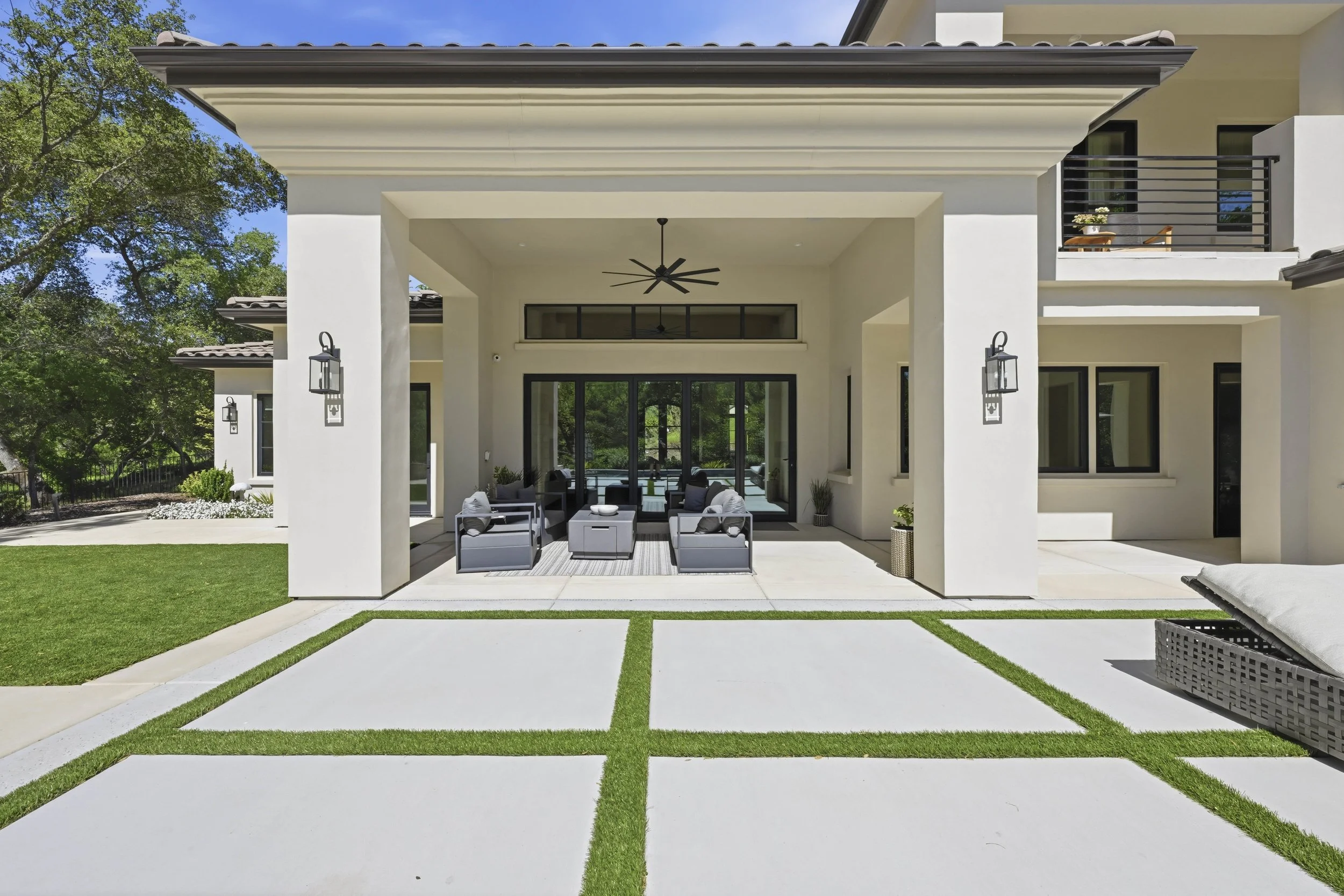 Modern house patio with outdoor furniture, white walls, black-framed windows, ceiling fans, and lush green trees in the background.