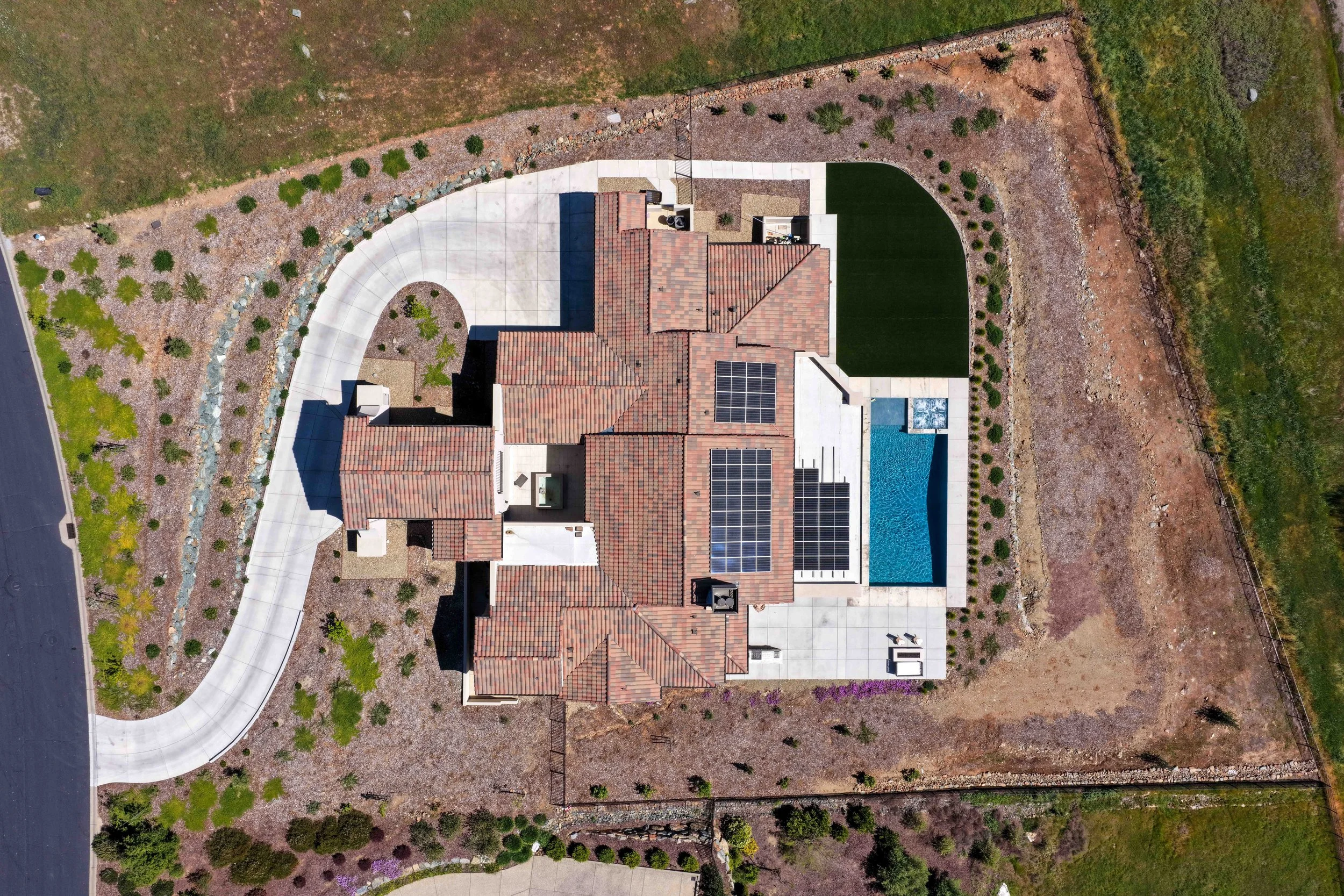 An aerial view of a large house with a red tile roof and a backyard with a swimming pool. The house is surrounded by a landscaped yard with trees, shrubbery, and a curved sidewalk on one side.
