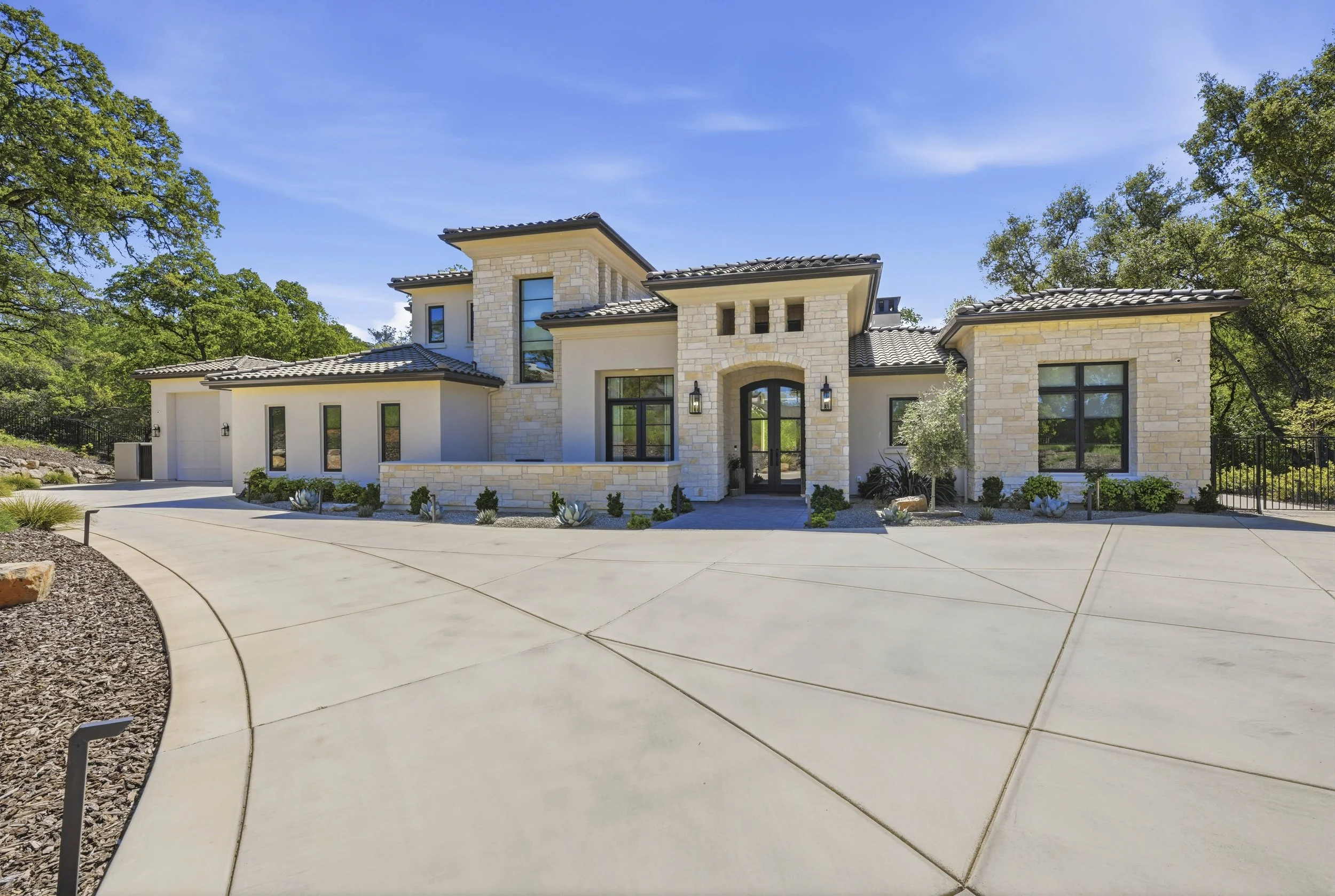 Modern house with stone and stucco exterior, large windows, attached garage, surrounded by greenery, under a clear blue sky.