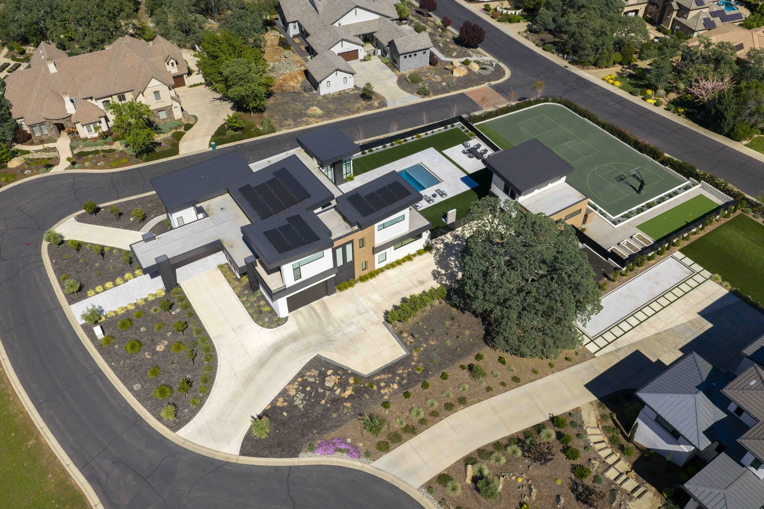 Aerial view of a modern residential neighborhood with houses, driveways, trees, and a residential community sports court featuring a basketball court and tennis court, surrounded by manicured lawns and landscaping.