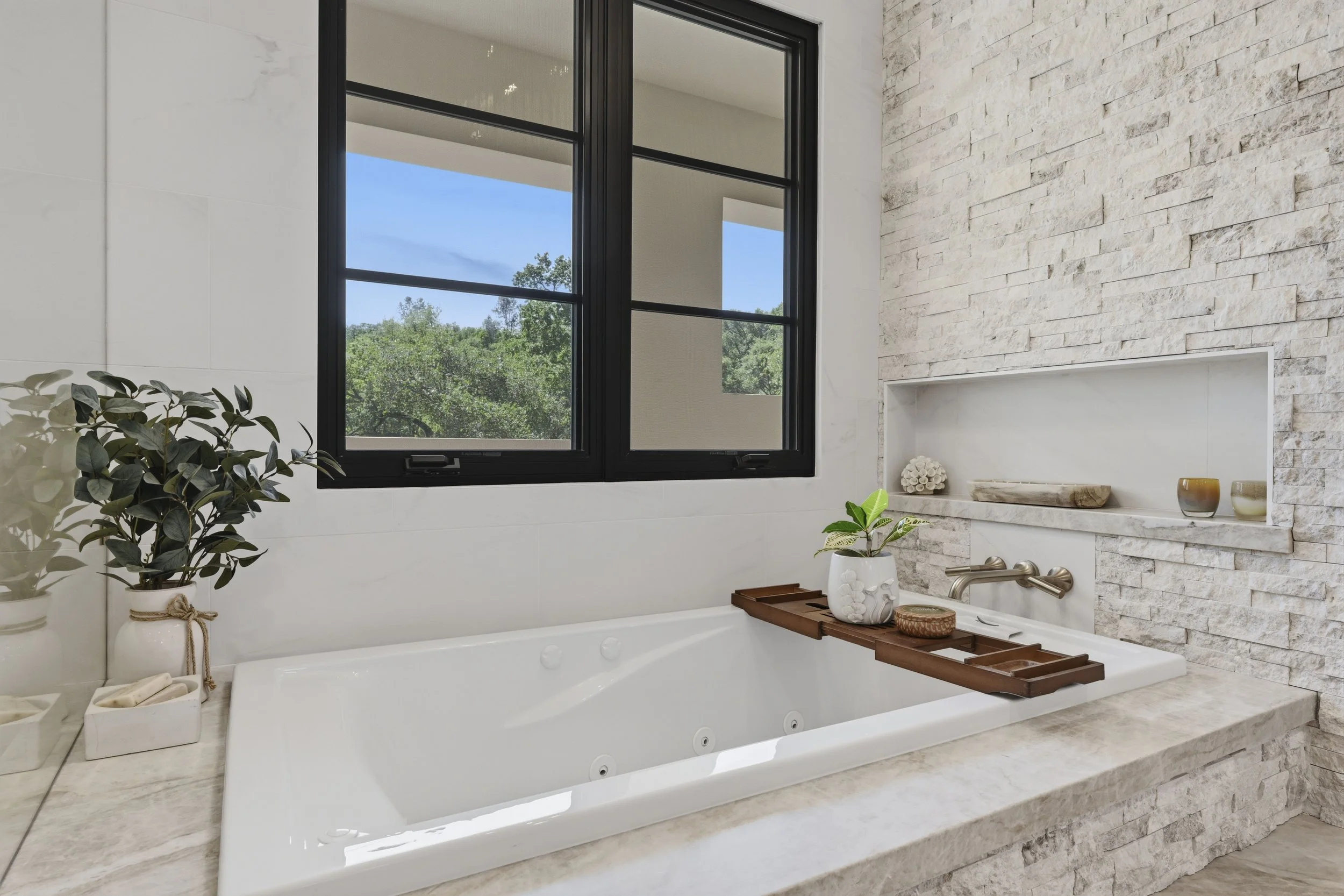 A bathroom with a bathtub under a large window showing a view of green trees and a blue sky. The bathroom has a stone wall, a white wall, and a small shelf with decorative items. There are potted plants nearby.